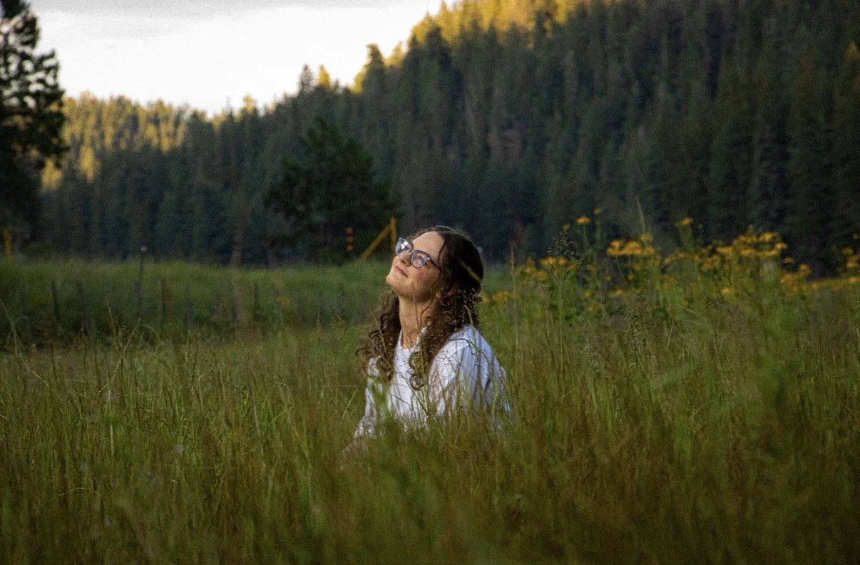 A woman with curly hair, glasses, and a white shirt sitting in a grassy field surrounded by yellow flowers and trees, with her eyes closed and head tilted upward.