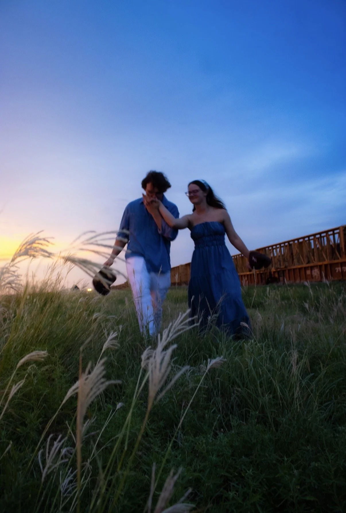A young man and woman walking through a grassy field at sunset, holding hands. The woman is wearing a strapless blue dress and glasses, while the man has curly hair and is wearing a blue shirt and white pants. There is a wooden fence in the background and tall grass in the foreground.