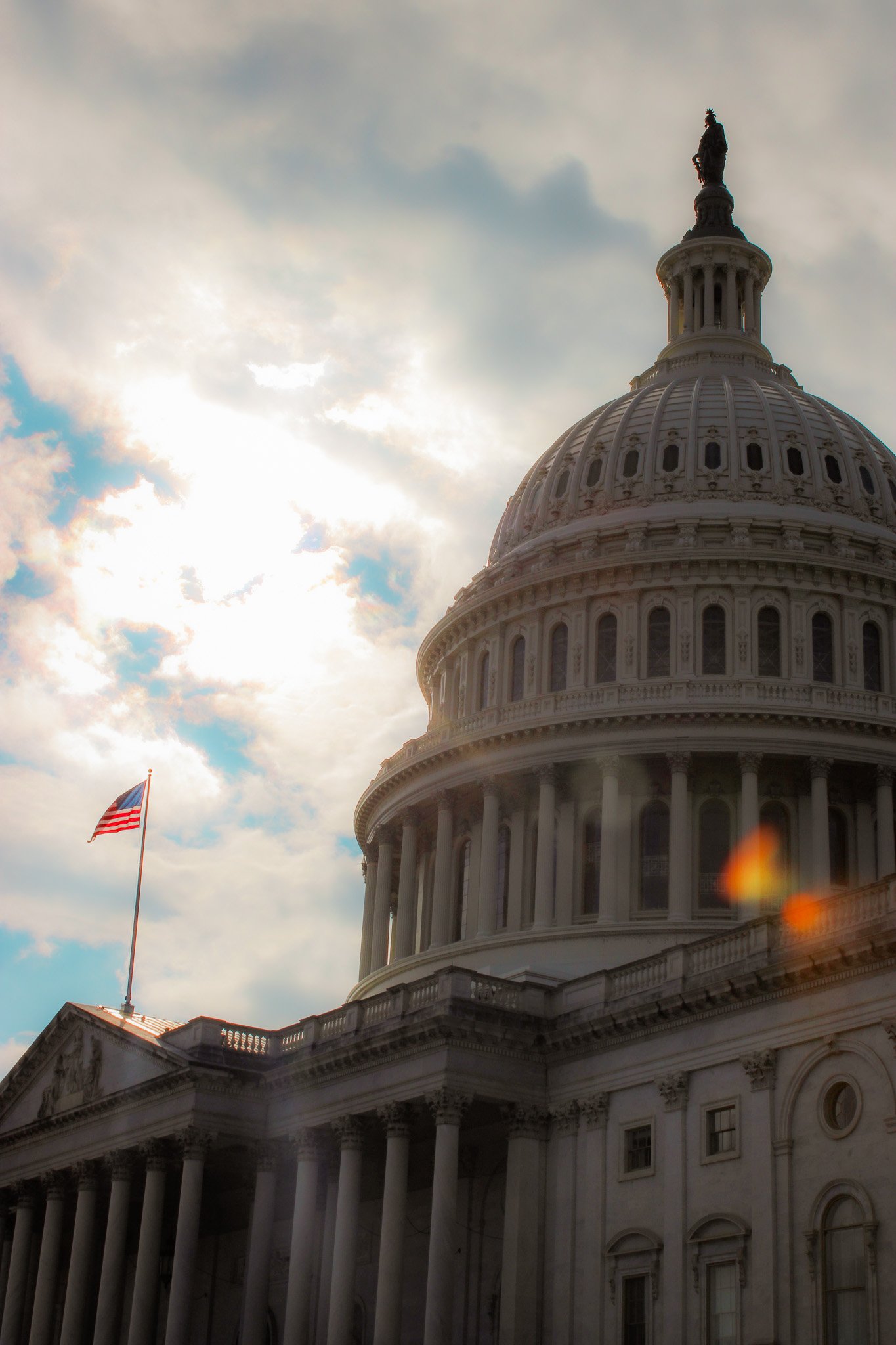 The United States Capitol building with the American flag flying in front. The sky is partly cloudy with the sun shining behind the clouds.