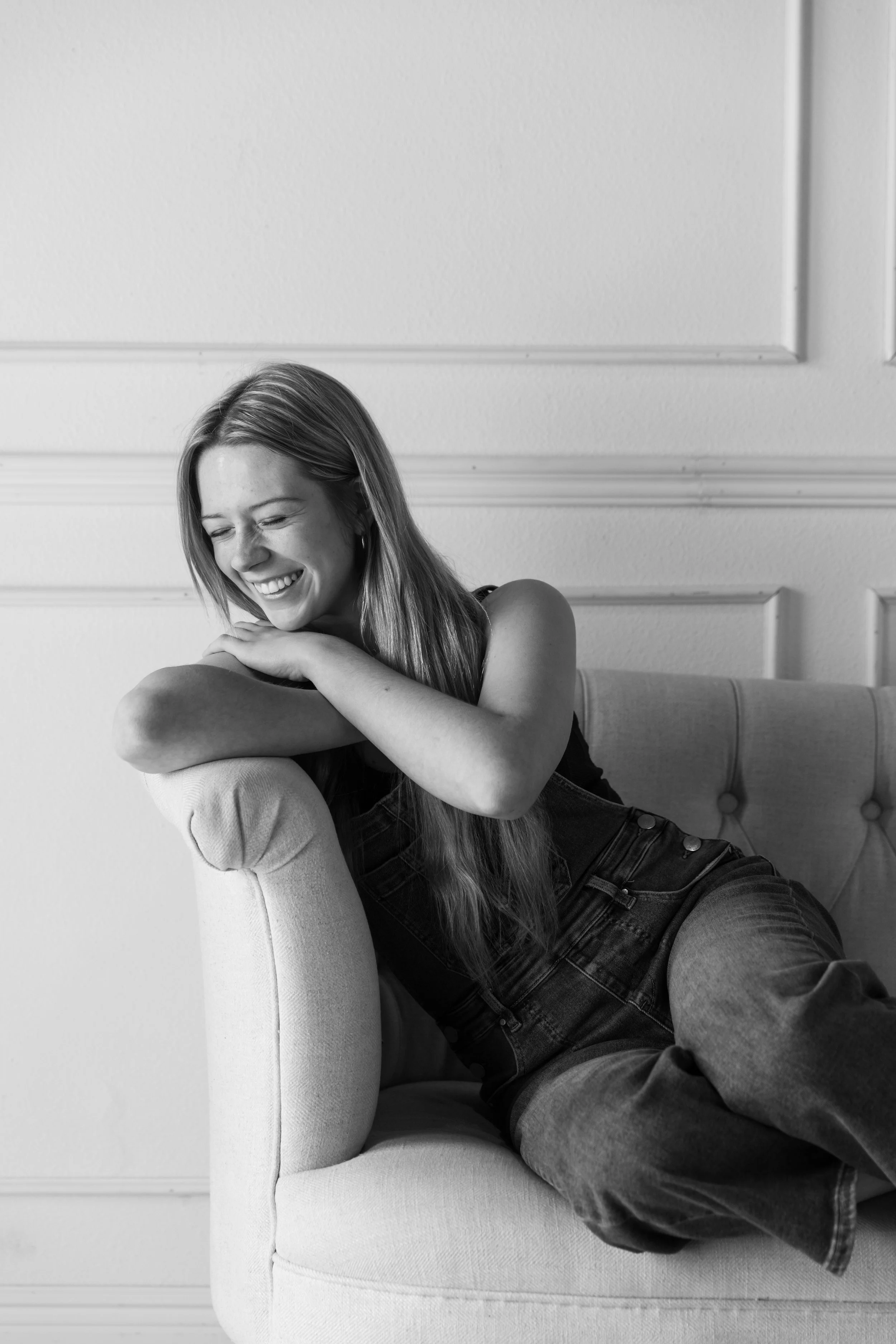A woman with long hair smiling and laughing, sitting on a sofa, leaning against the armrest, with her arms crossed over her knees, in a black and white photo.