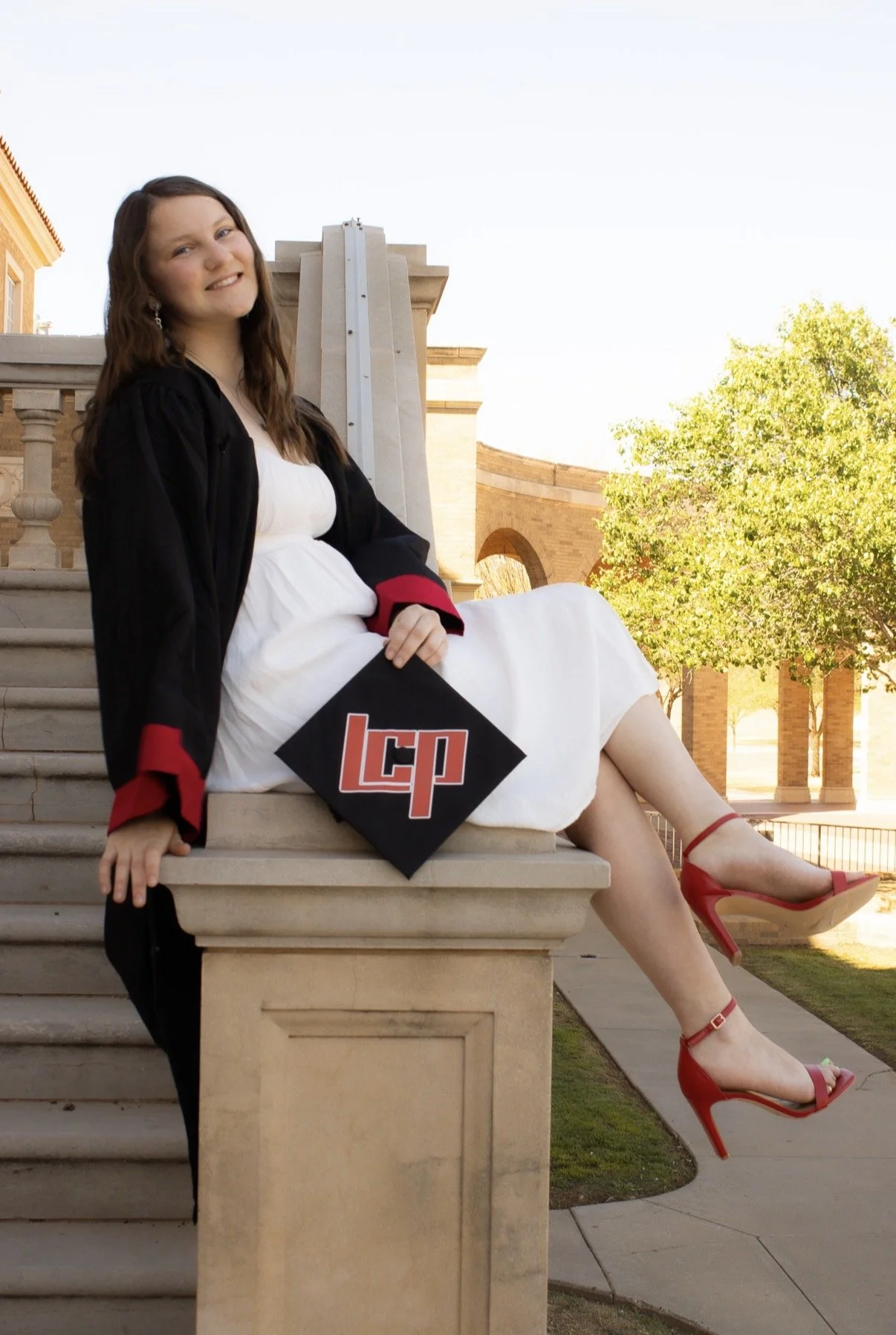 A young woman in a white dress and red high heels, wearing a black graduation gown with red trim, sitting on outdoor steps holding a black graduation cap with red and white initials 'LCP' in white and red. She is smiling and looking at the camera, with a university building and green trees in the background.