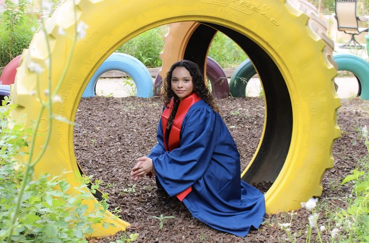 A young girl in a graduation gown kneeling inside a large yellow tire in an outdoor setting decorated with colorful tires and greenery.