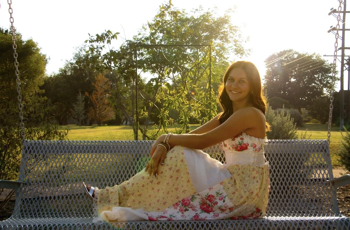 A smiling woman with long brown hair sitting on a white park bench swing during sunset in a park with trees and greenery.