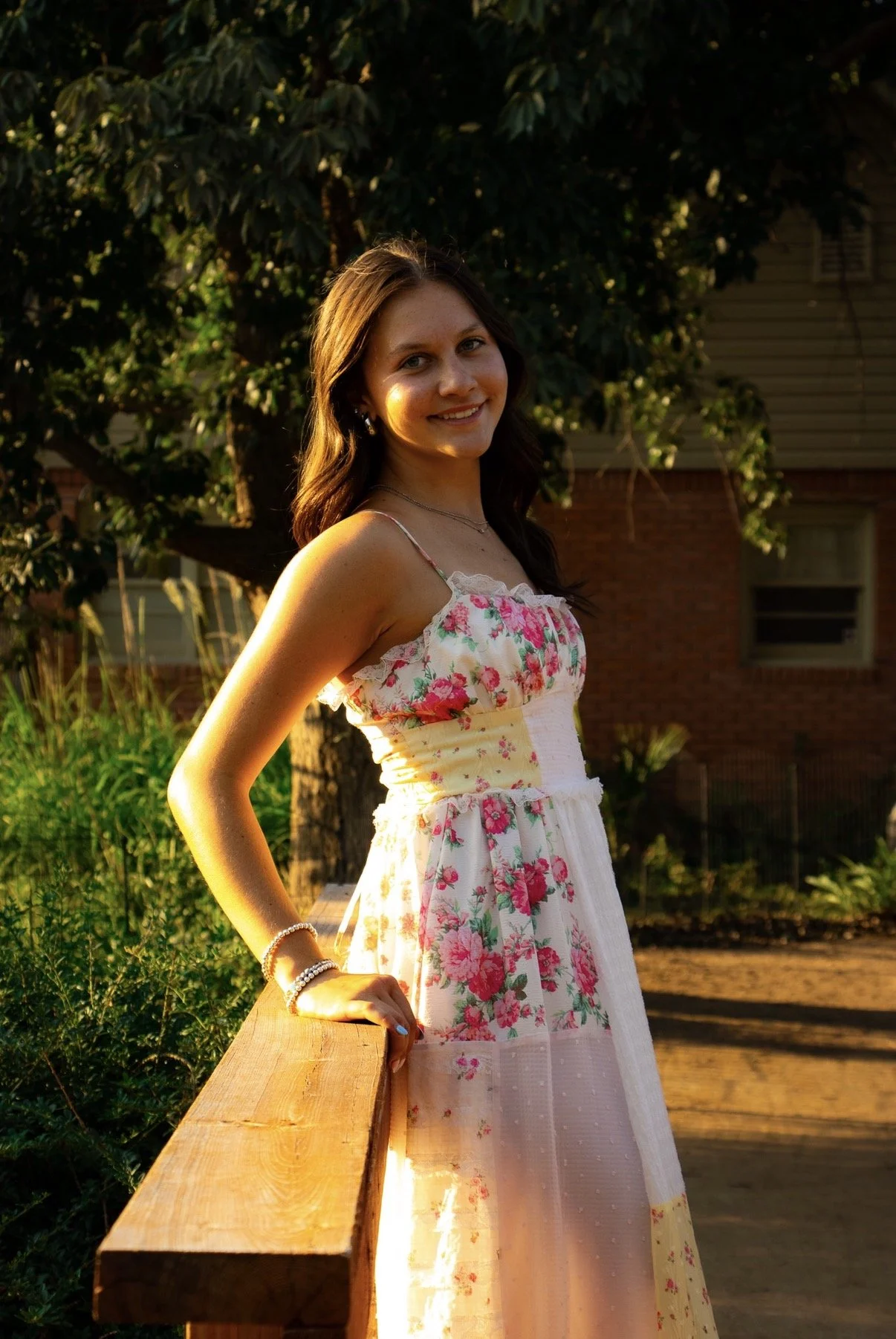 A young woman with long brown hair smiling, standing outdoors with a wooden railing and green trees in the background during sunset.