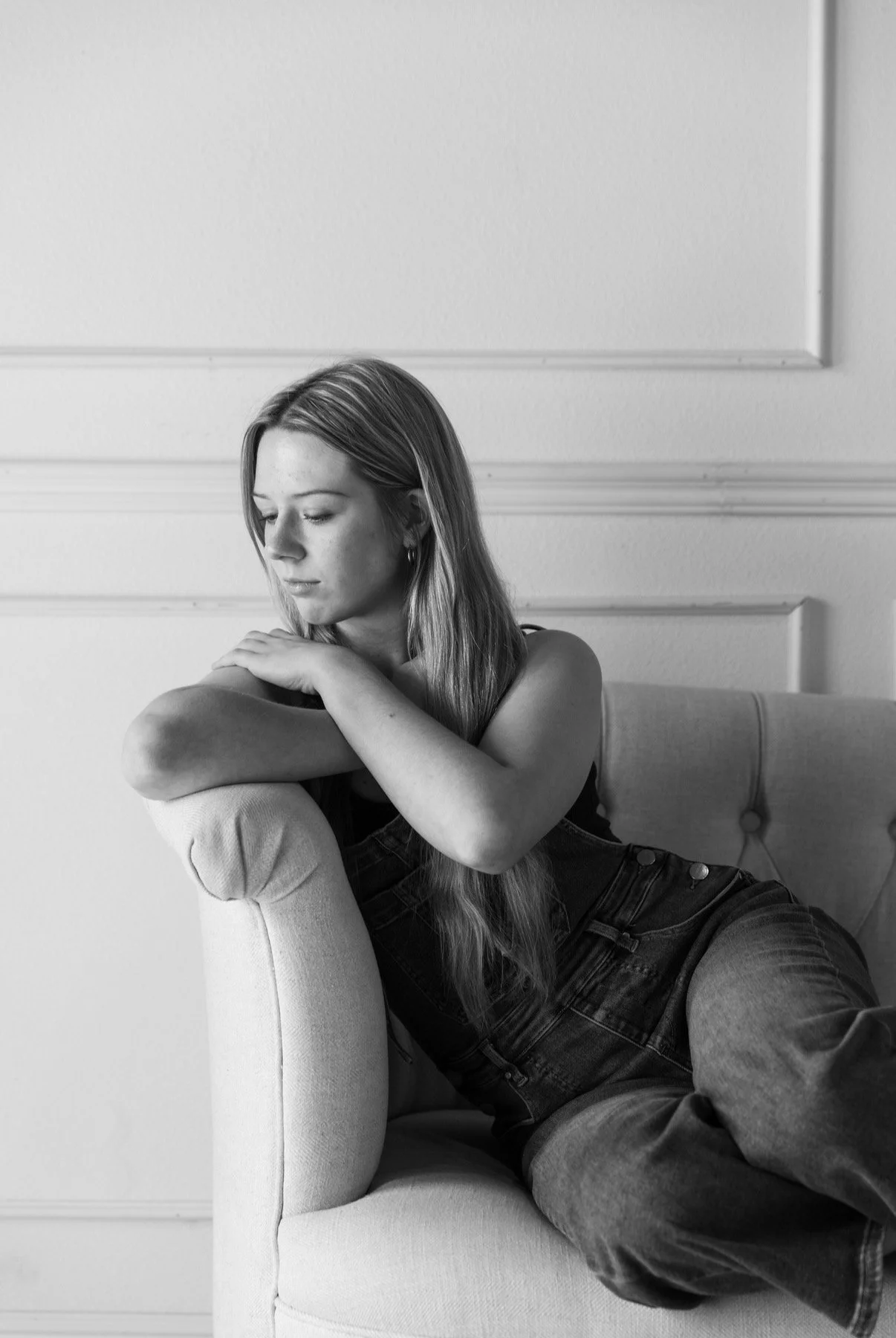 Black and white photo of a young woman with long hair, sitting on a sofa with her arm resting on the back, looking down thoughtfully.