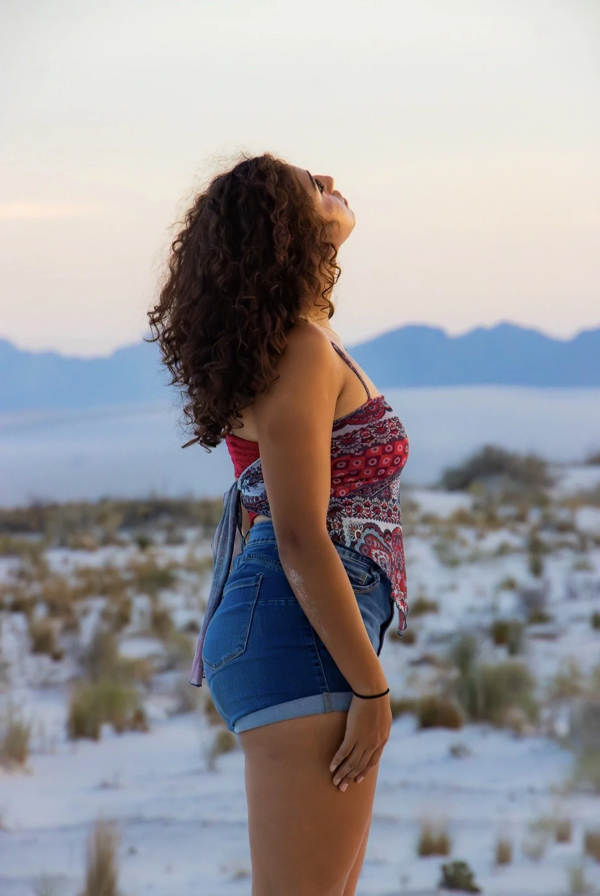 A woman with curly hair standing outdoors in a desert landscape during sunset, wearing a colorful tank top and denim shorts, looking up at the sky.