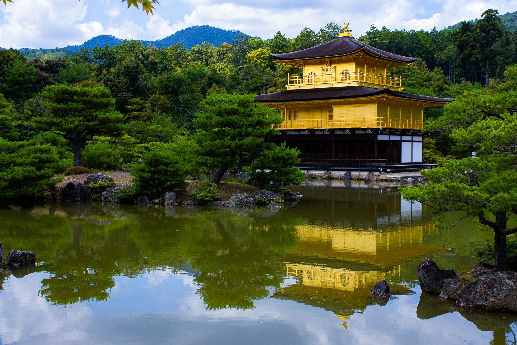 A traditional Japanese building with a dark, curved roof, painted yellow, located among lush green trees and reflected in a calm pond in a scenic mountain landscape.