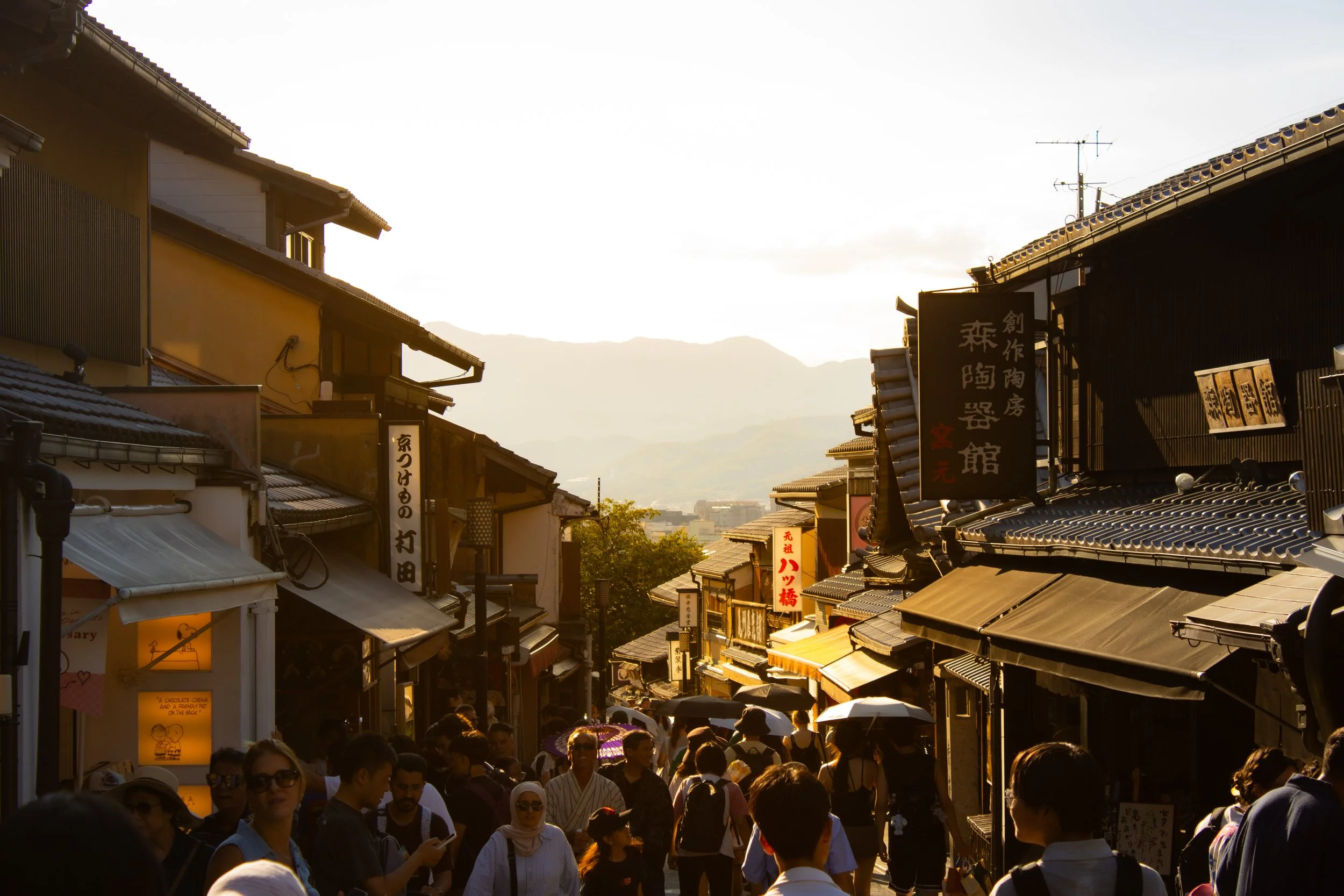 Crowded street scene during sunset with people walking and shopping, traditional Japanese buildings on either side, and signs in Japanese characters.