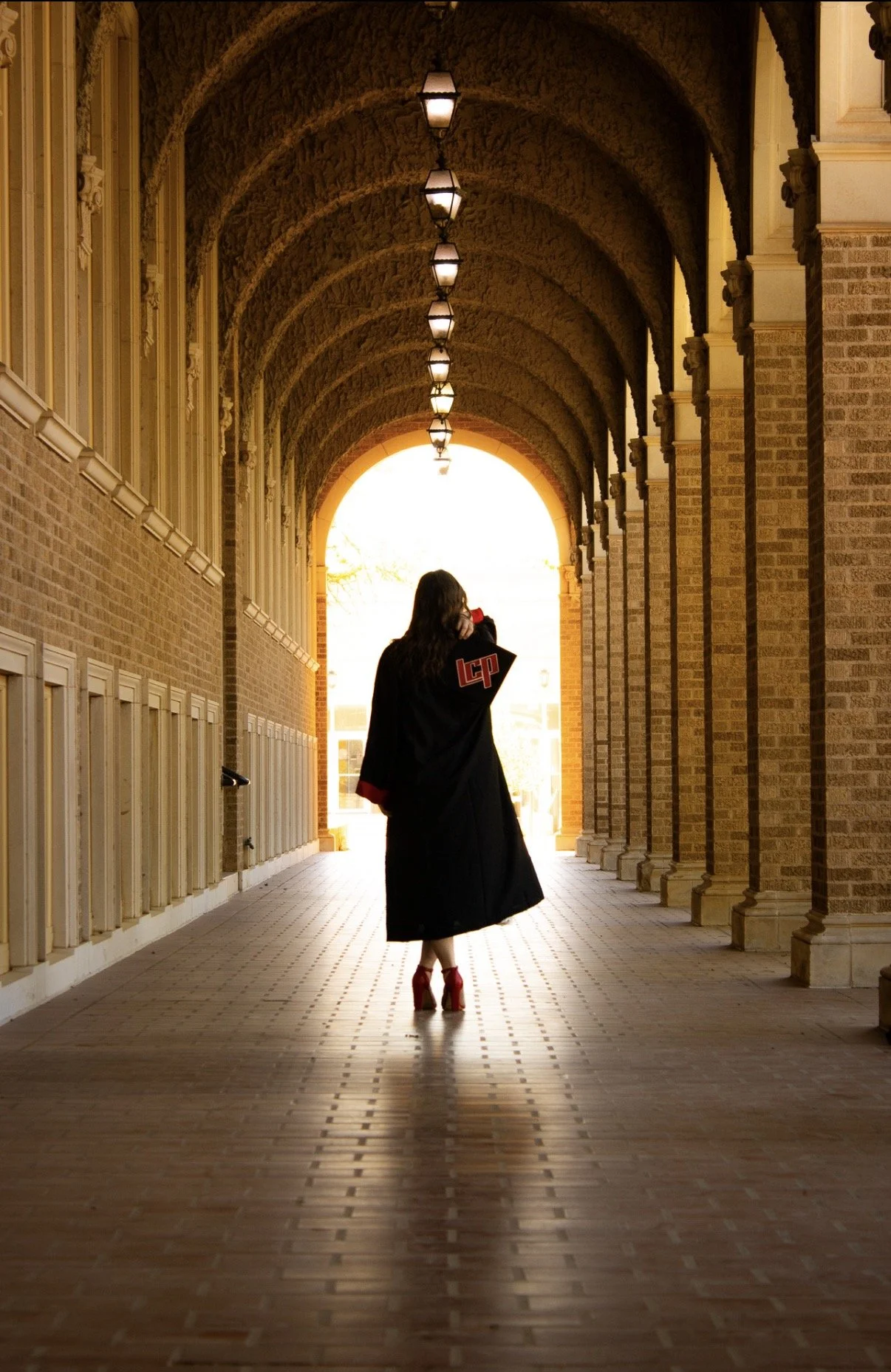 A woman in a black graduation gown and red high heels walking through a brick archway corridor with hanging lights, backlit by sunlight.