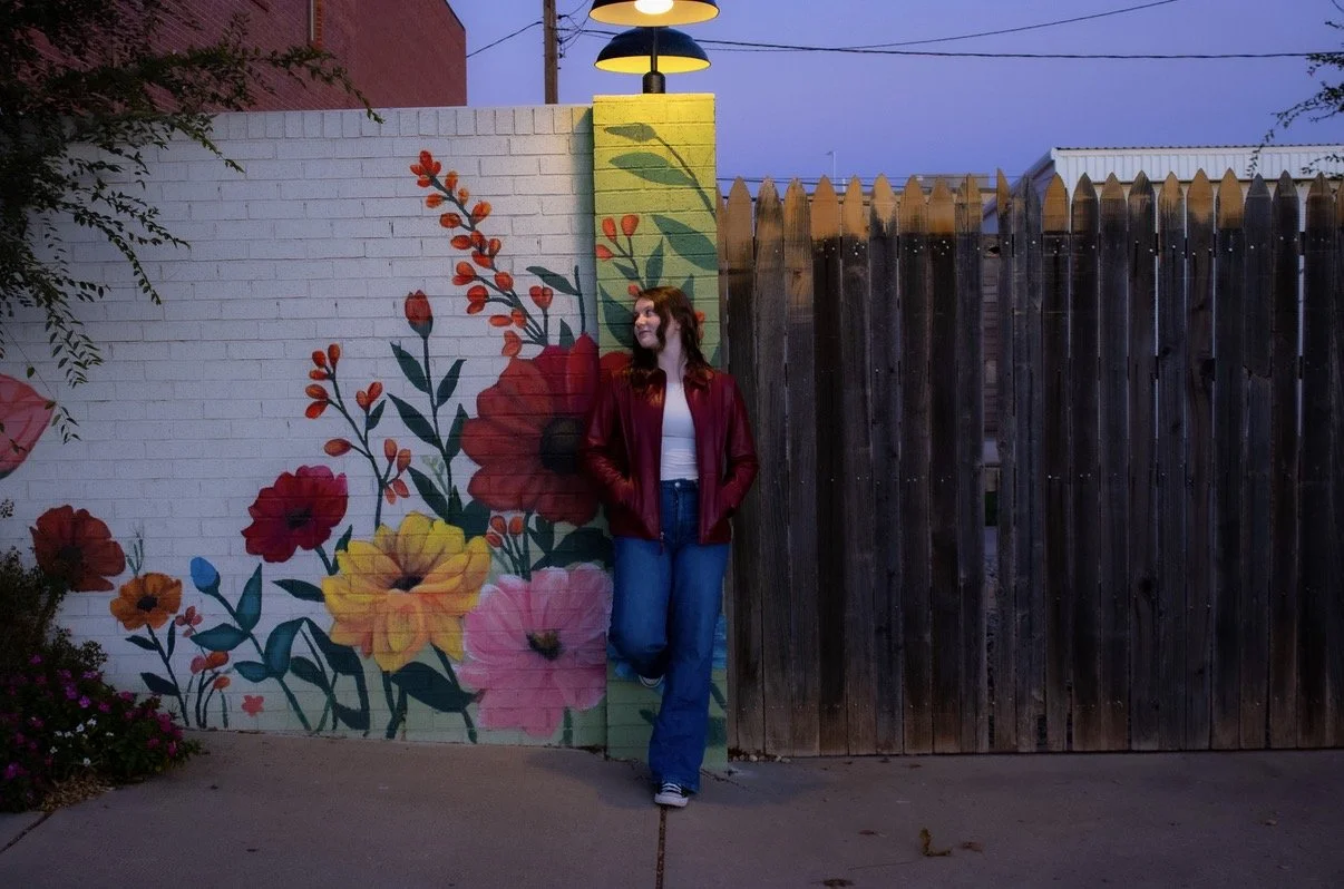 A woman with reddish-brown hair wearing a red leather jacket, white top, and jeans stands against a mural of colorful flowers painted on a brick wall, with a wooden fence to her right, during dusk or early evening.