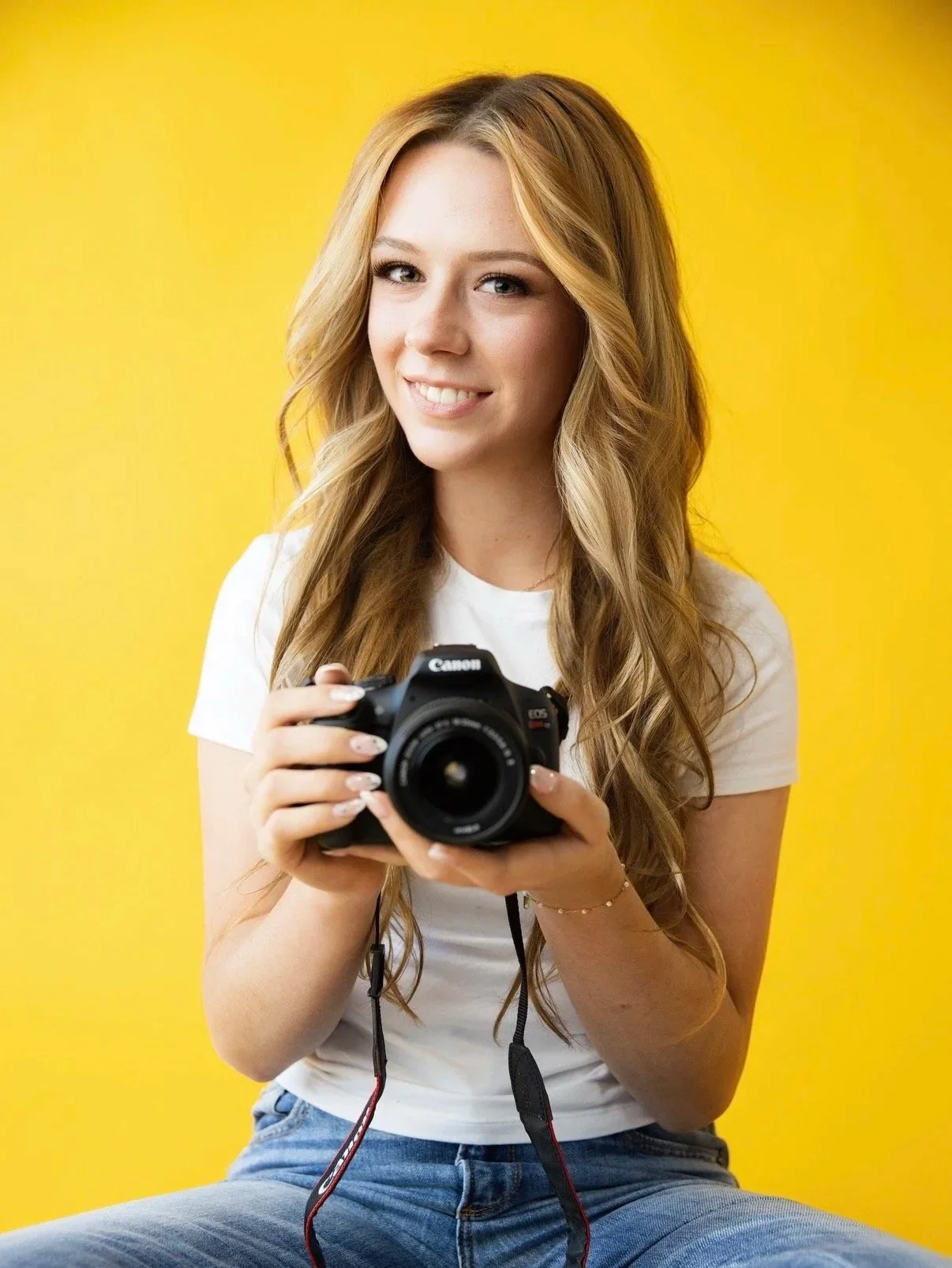 A young woman with long blonde hair, smiling, sitting against a yellow background, holding a Canon DSLR camera.