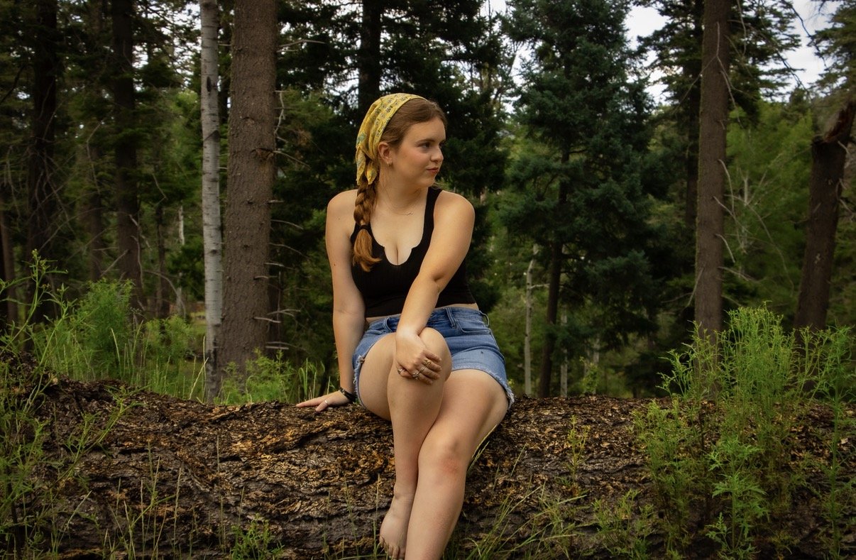 A young woman with red hair, wearing a yellow bandana, black tank top, and denim shorts, sitting on a fallen tree trunk in a forest with tall trees and green foliage.