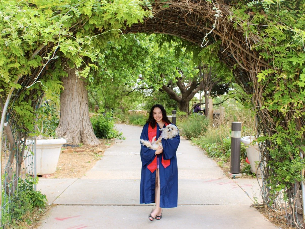 A young woman in a graduation gown and stole is smiling and holding a small dog while standing on a sunny sidewalk under a leafy archway.