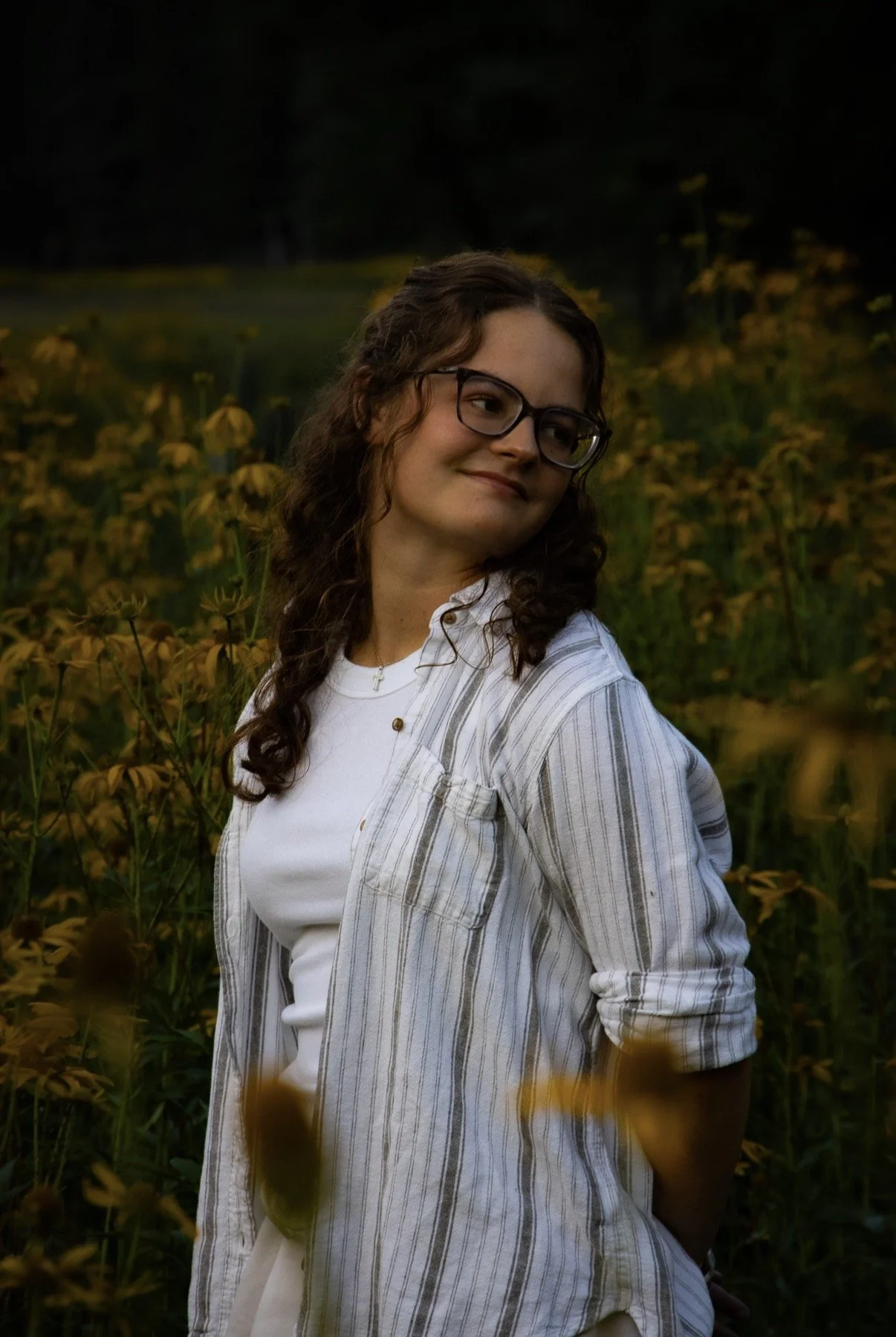 A young woman with long curly brown hair and glasses standing in a field of yellow flowers at dusk, wearing a white t-shirt and a striped shirt.