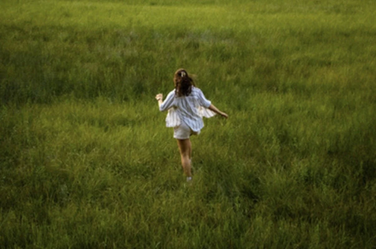 A girl running through a lush green field with her back facing the camera.