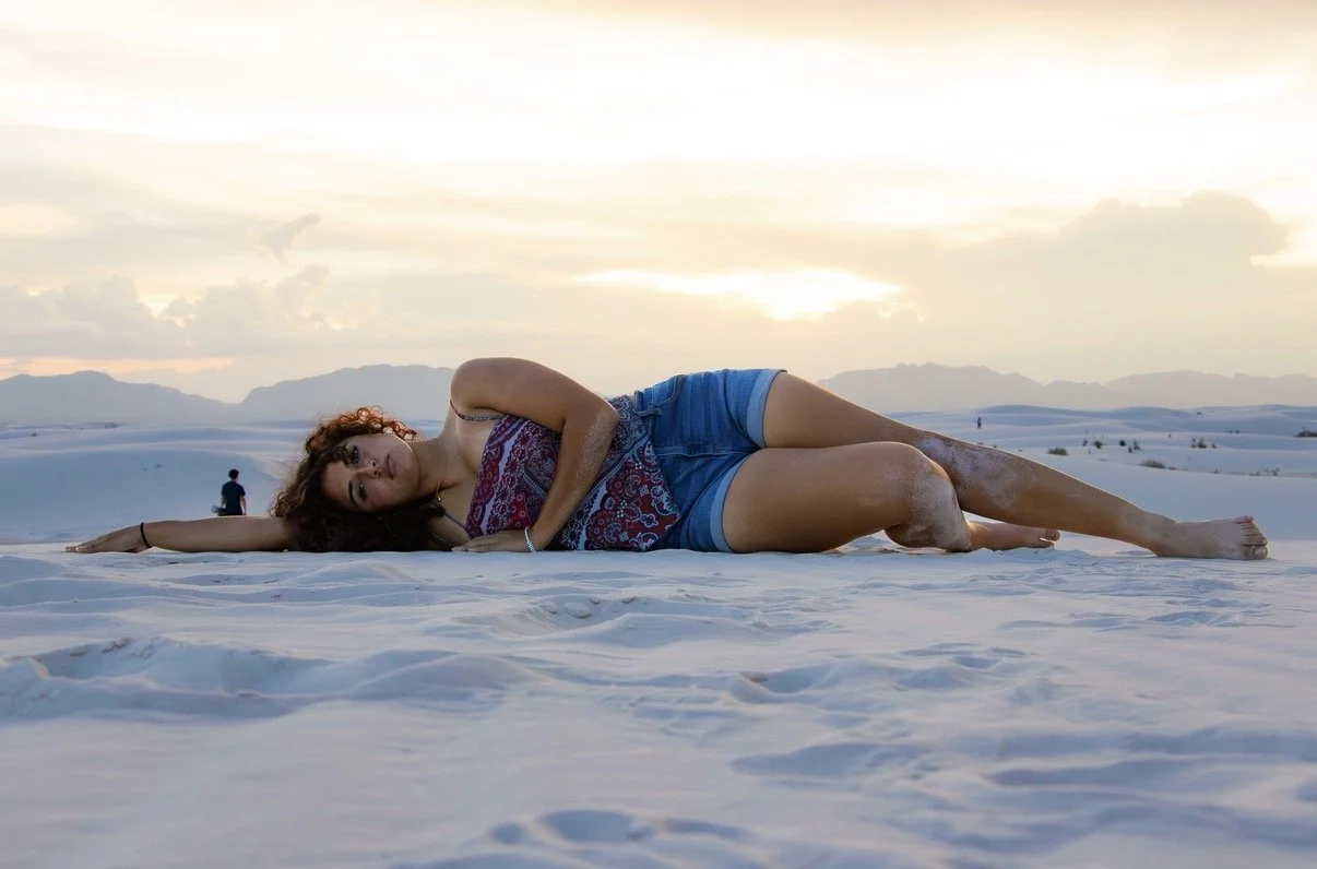 Woman lying on sand in a desert, wearing a patterned top and denim shorts, during sunset.