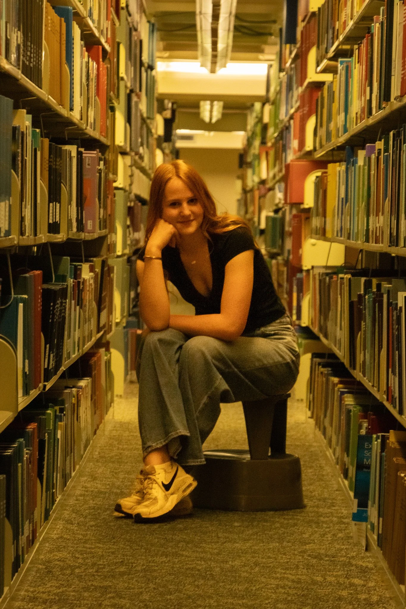 A young woman with red hair sitting on a stool in a library aisle, surrounded by bookshelves filled with books, smiling and resting her chin on her hand, wearing a black top, gray pants, and yellow sneakers.