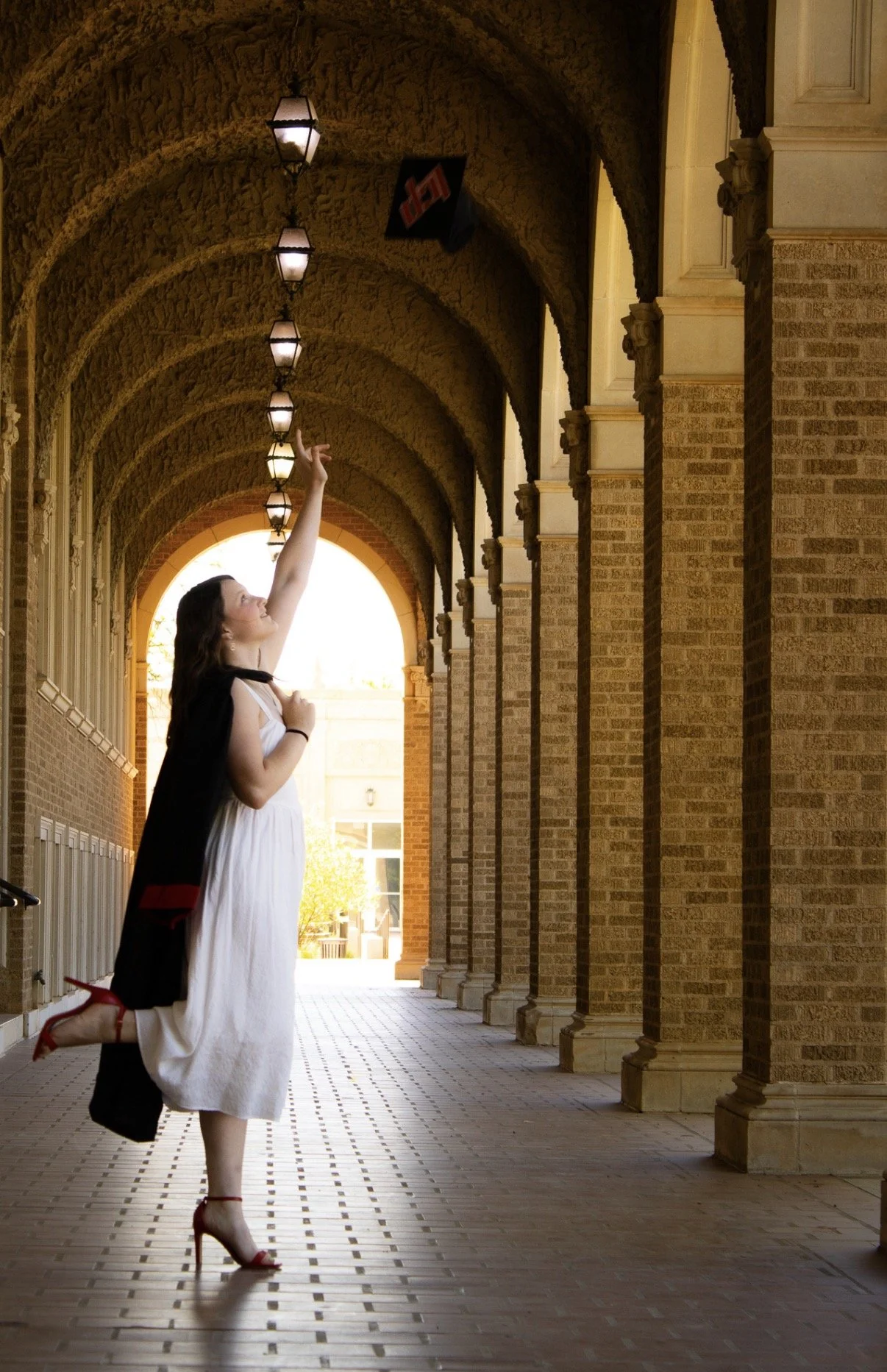 A woman kneeling on one leg, dressed in a white dress and red heels, lifts her arm to touch hanging lanterns in a covered brick walkway, with an arched opening in the background.