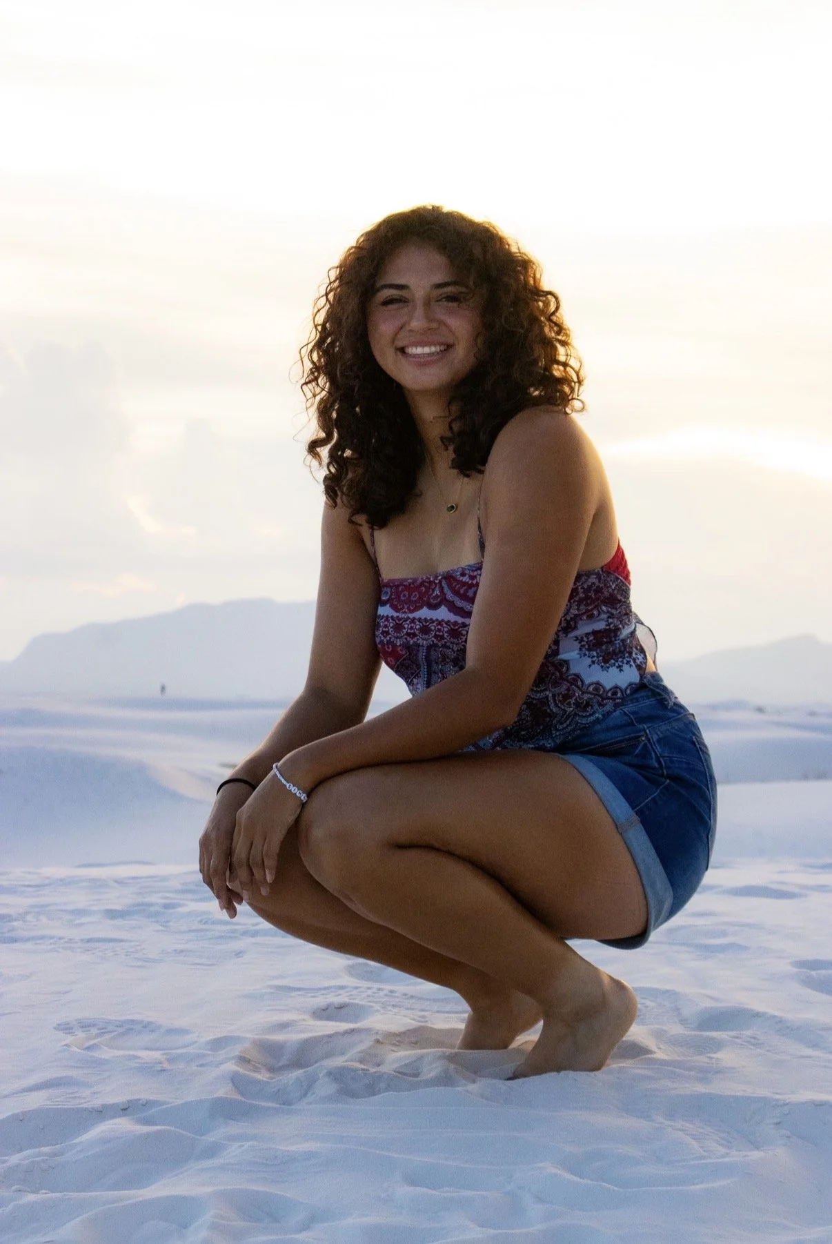 Young woman with curly hair smiling, squatting on white sandy ground, wearing a colorful sleeveless top and denim shorts, with mountains in the distance and a bright sky.