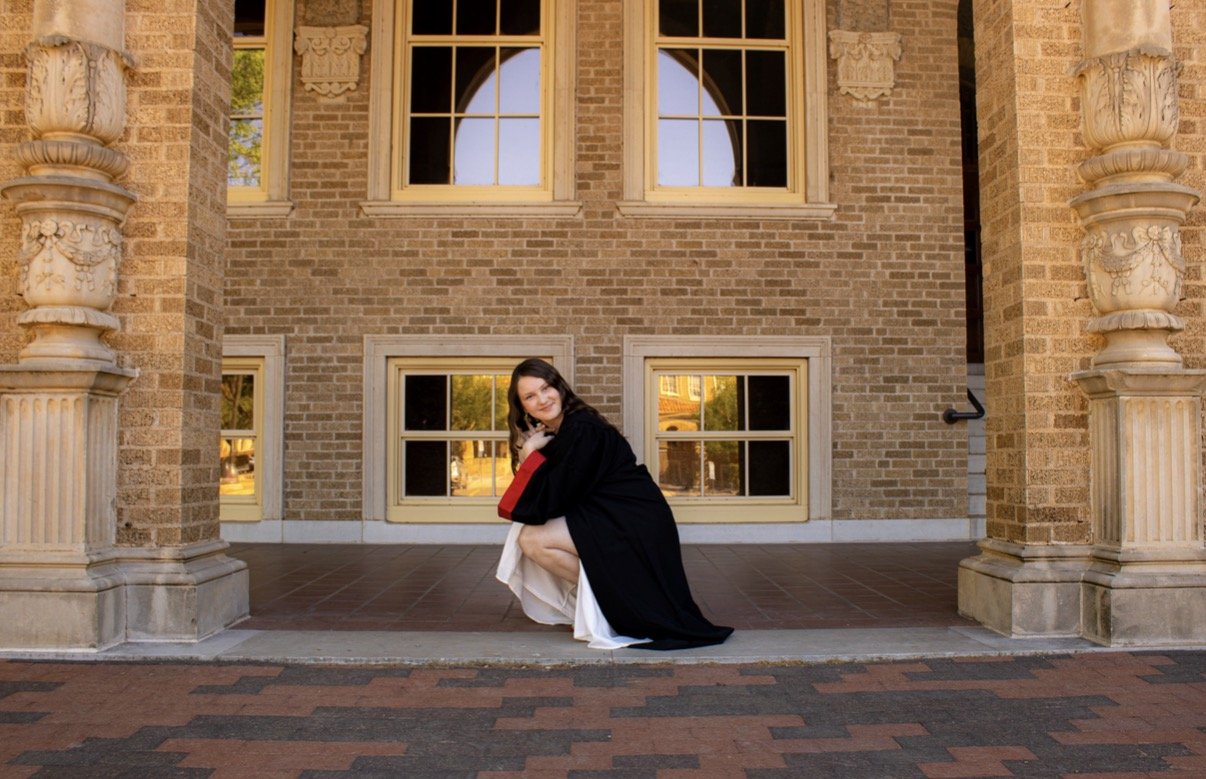 A woman in graduation attire, kneeling on a sidewalk in front of a brick building with large windows and ornate columns.