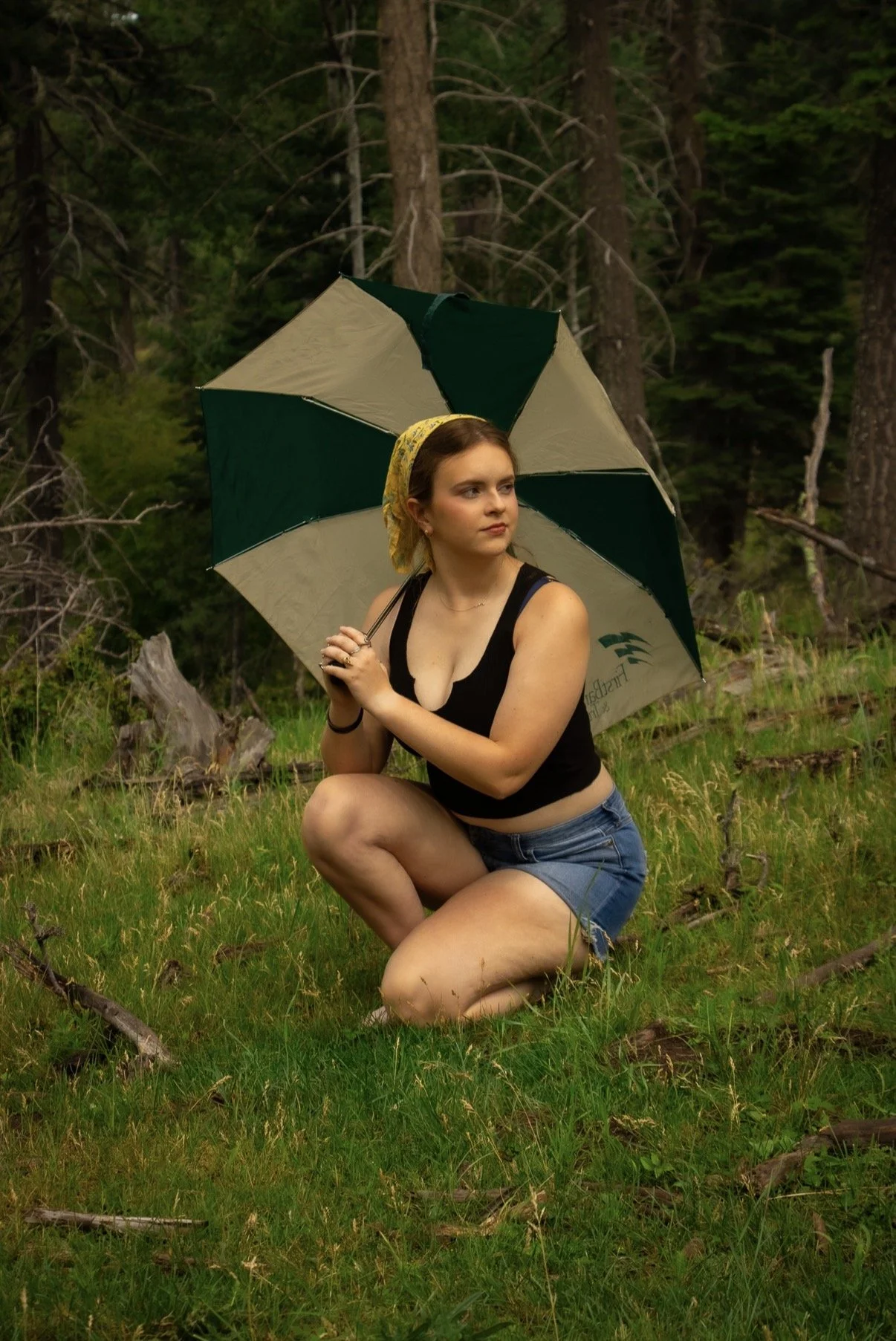 A woman crouching on the grass in a forest, holding a large green and beige umbrella, wearing a black tank top, denim shorts, and a yellow headscarf.