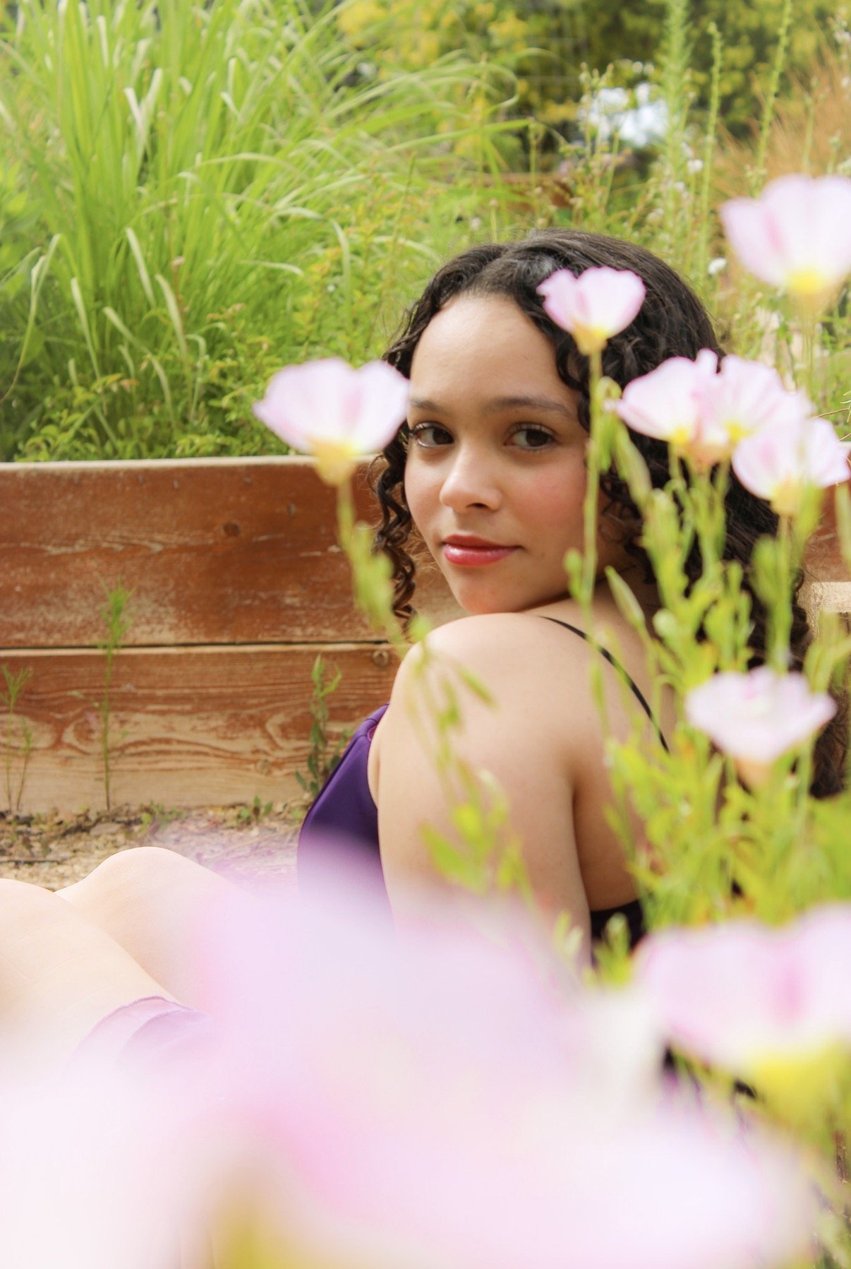 A young woman with curly dark hair and light skin sitting outdoors near a wooden planter box, surrounded by pink and white flowers and green foliage, looking at the camera with a slight smile.
