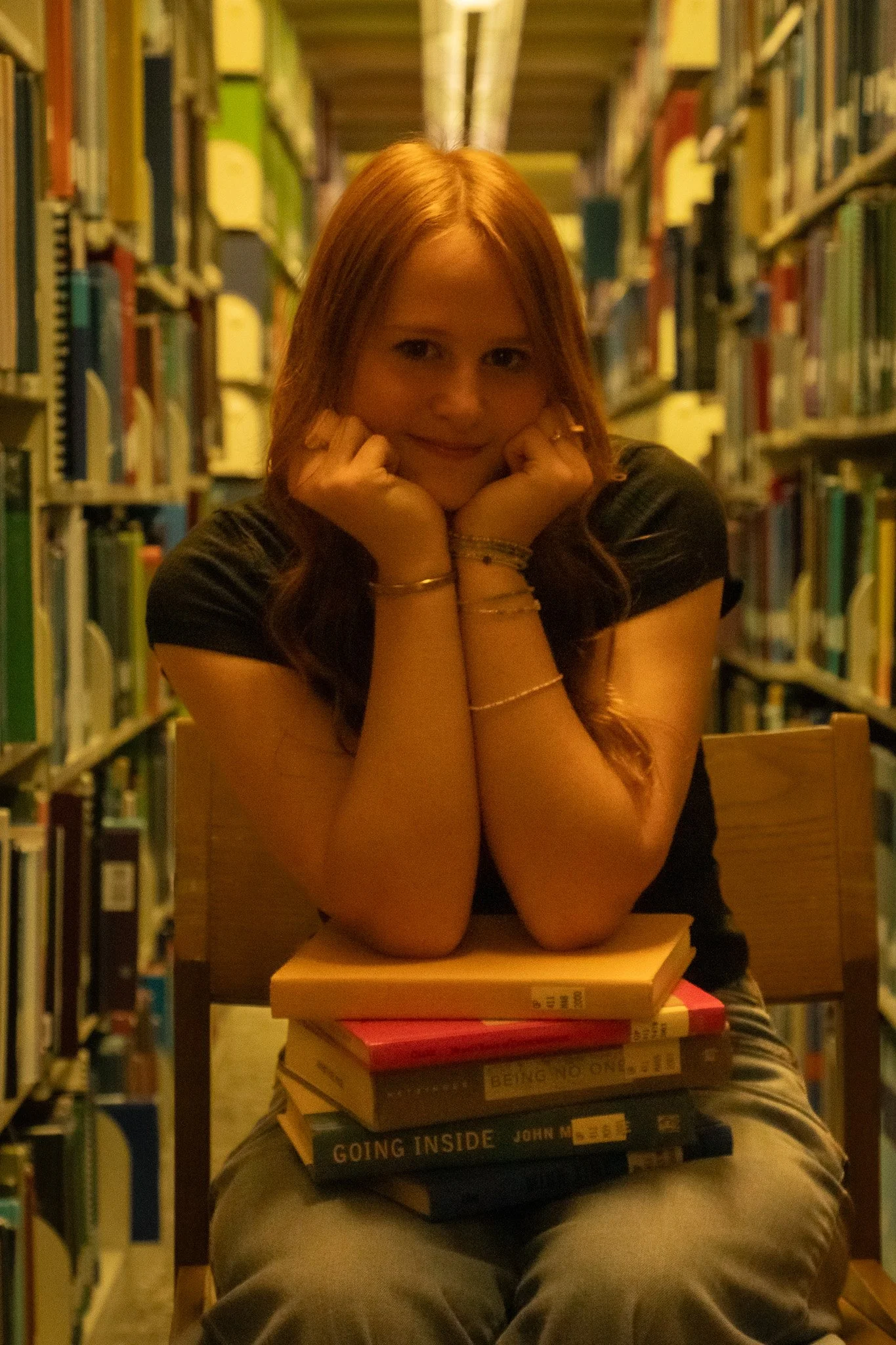 A young woman with red hair sitting in a library, resting her chin on her hands, with a stack of books on her lap.