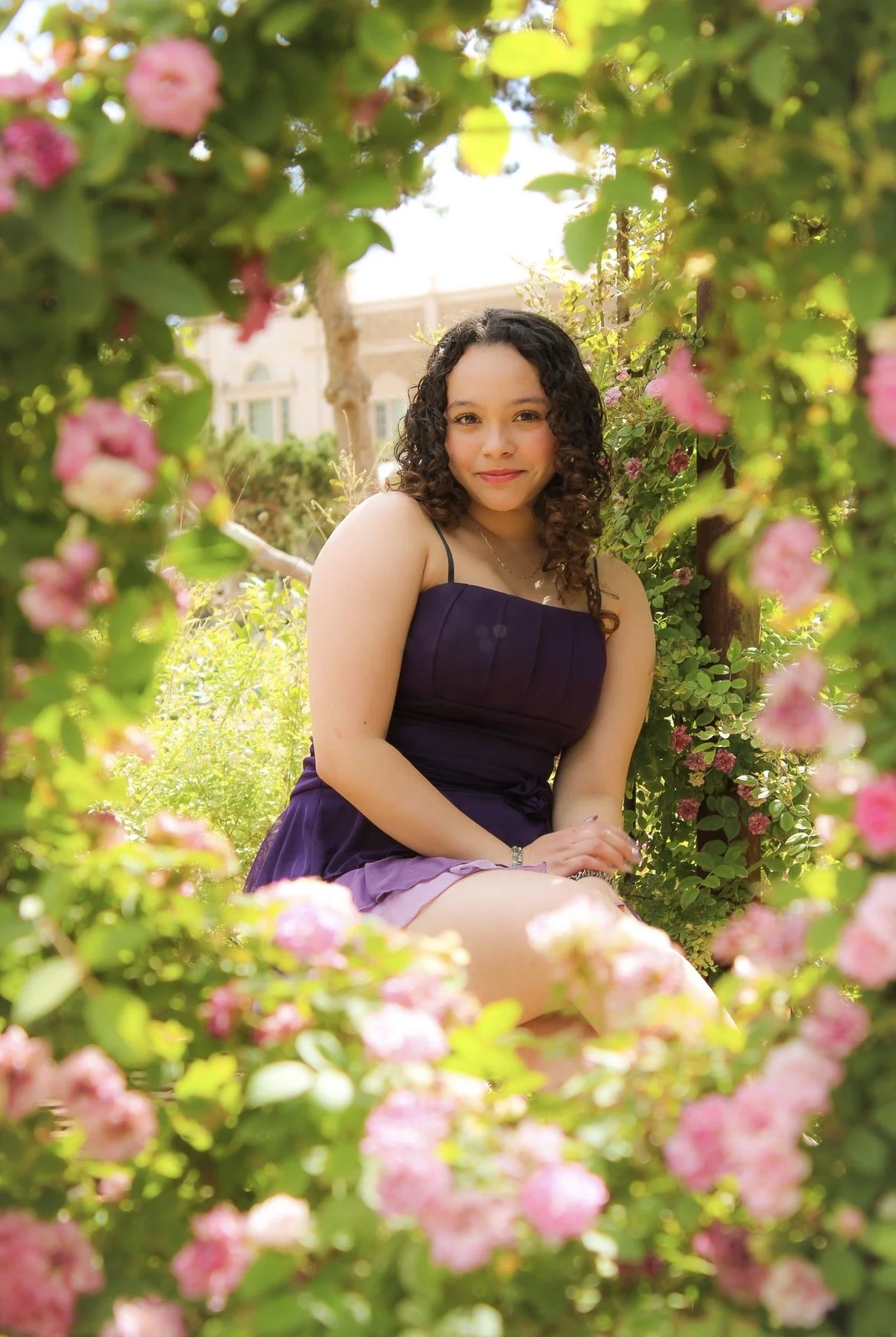 Young woman sitting outdoors among pink flowering bushes, smiling at the camera, wearing a dark purple dress.