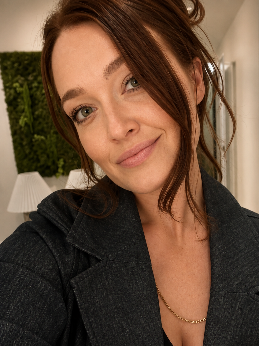 Close-up of a Megan Willerford with auburn hair, green eyes, and light makeup, smiling slightly, in an indoor setting with a white lampshade and greenery in the background.