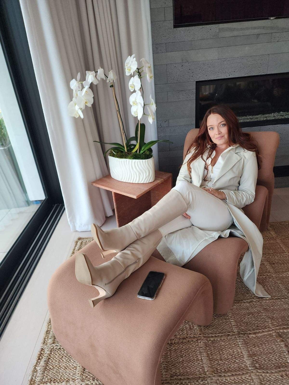 A woman with long, wavy, brown hair, wearing a white blazer and matching white knee-high boots, sitting on a pink chaise lounge in a modern living room. She is relaxed, with one leg crossed over the other, and a black smartphone resting on the lounge next to her. Behind her, there is a gray stone wall, a beige armchair, a wooden side table with a white orchid plant, white curtains, and large glass sliding doors leading outside.