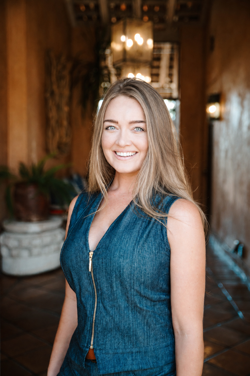 A smiling Megan Willerford with long blonde hair and blue eyes, wearing a sleeveless navy blue top with a zipper, standing indoors with warm lighting and wooden decor in the background.