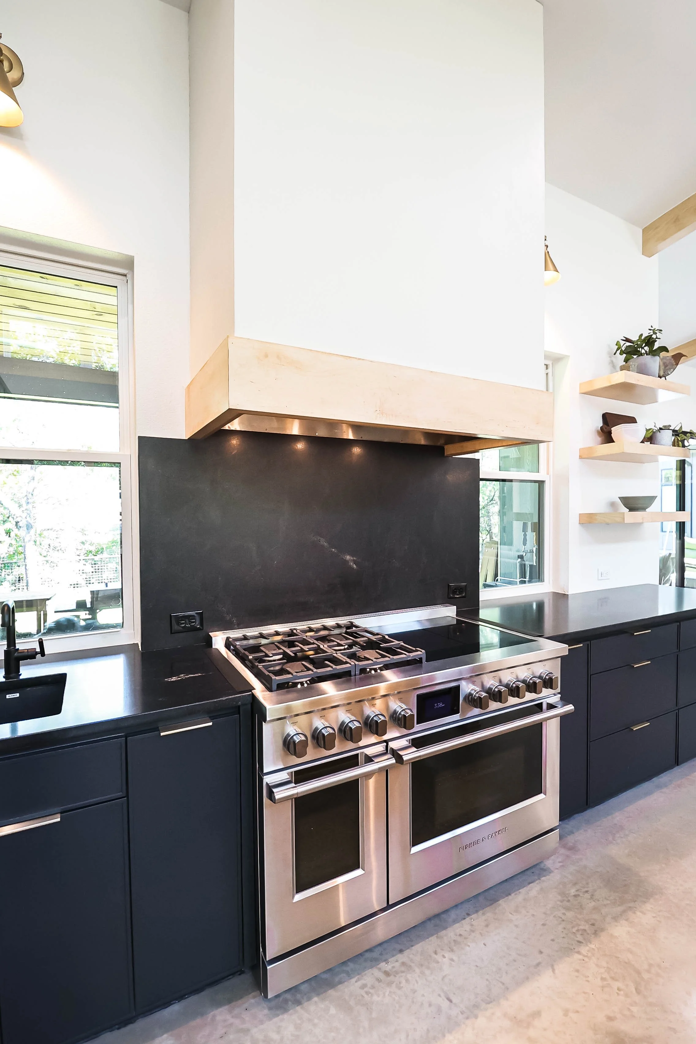 Modern kitchen with black cabinetry, stainless steel stove with multiple burners, black countertops, and open wooden shelves with potted plants and decorative bowls. Large windows provide natural light.