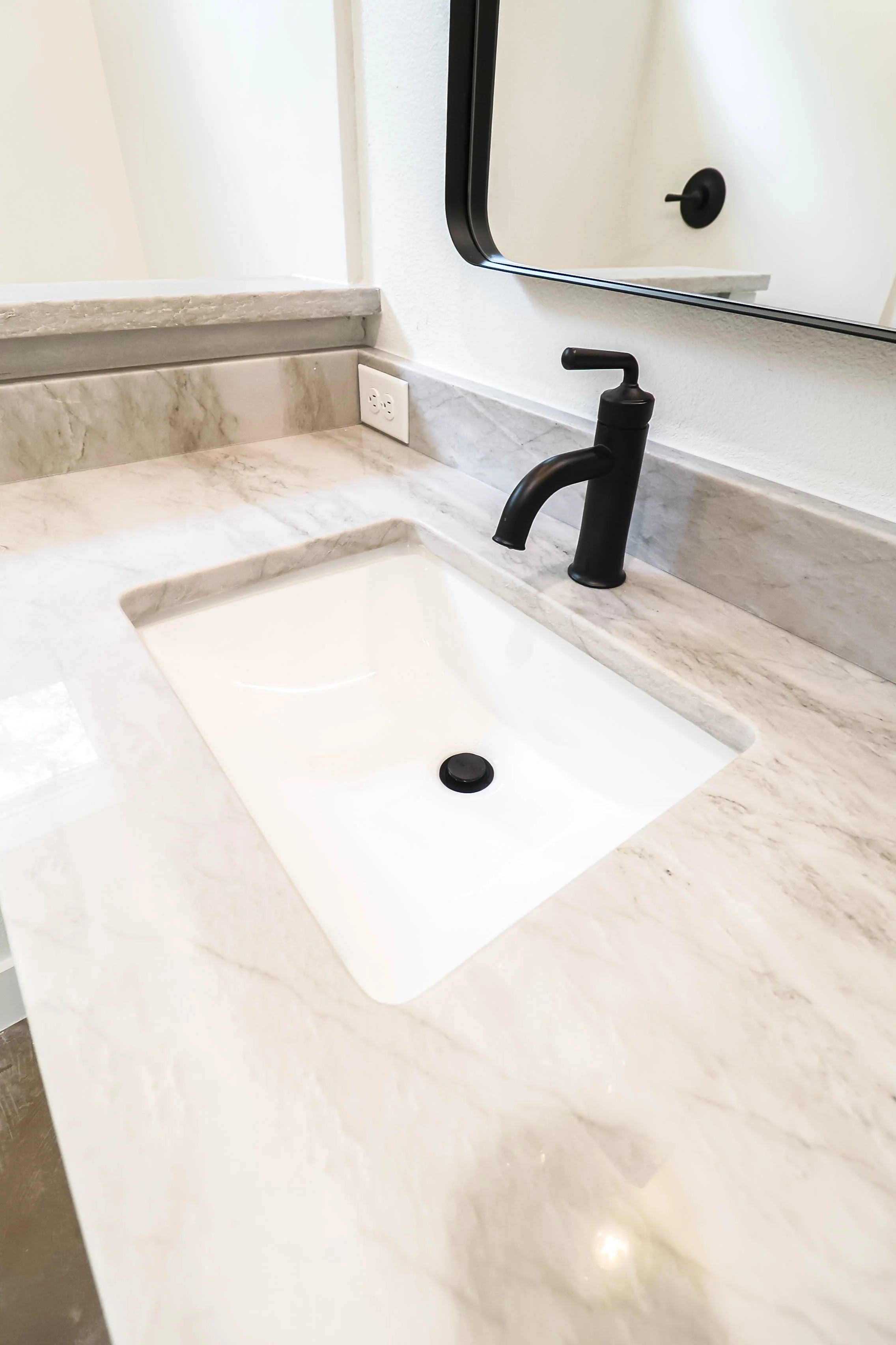 Close-up of a bathroom countertop with a black faucet, a mirror, and a wall outlet.