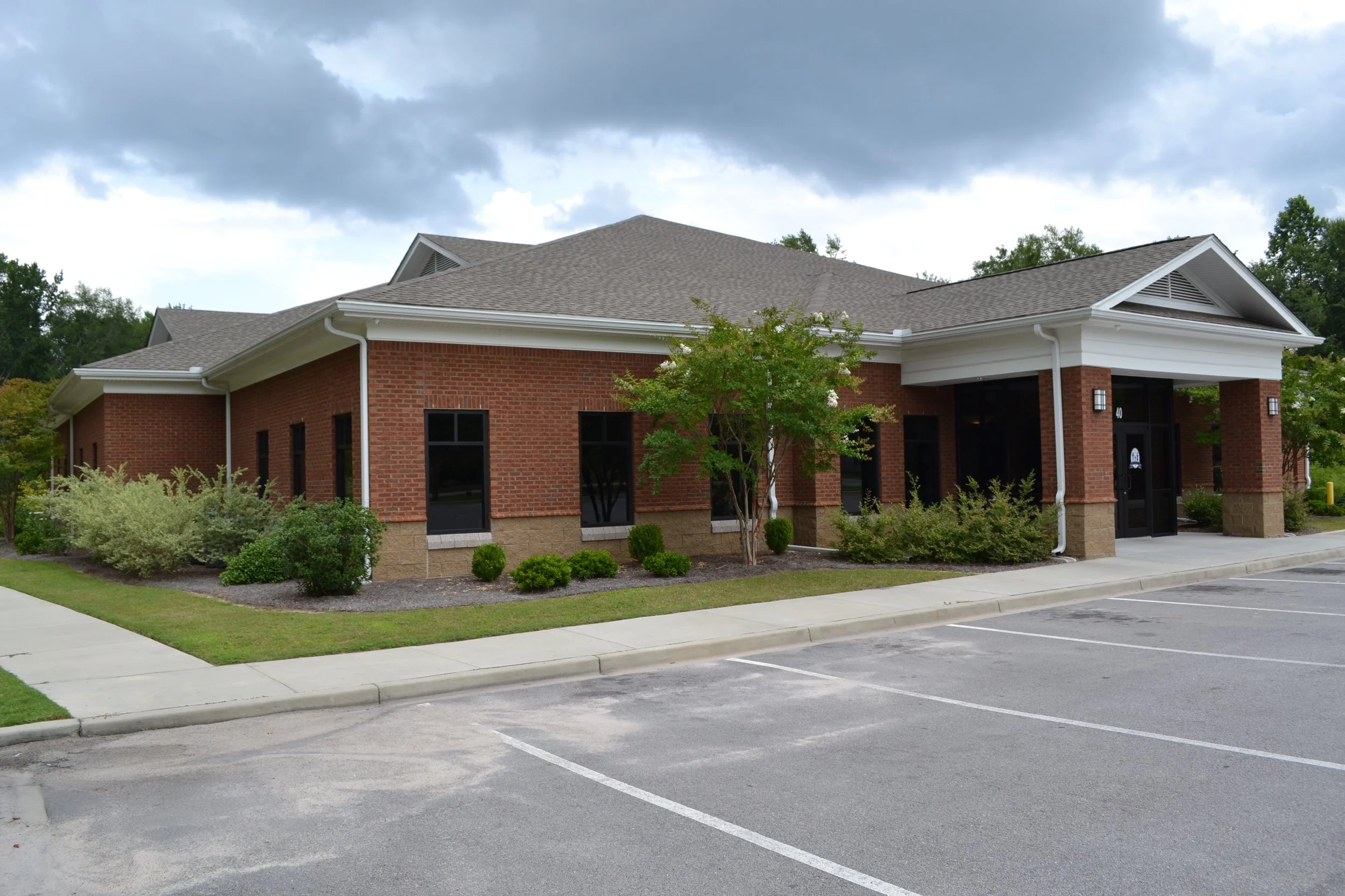 A brick building with white trim and a gabled roof, surrounded by greenery, with a parking lot with empty spaces in the foreground.