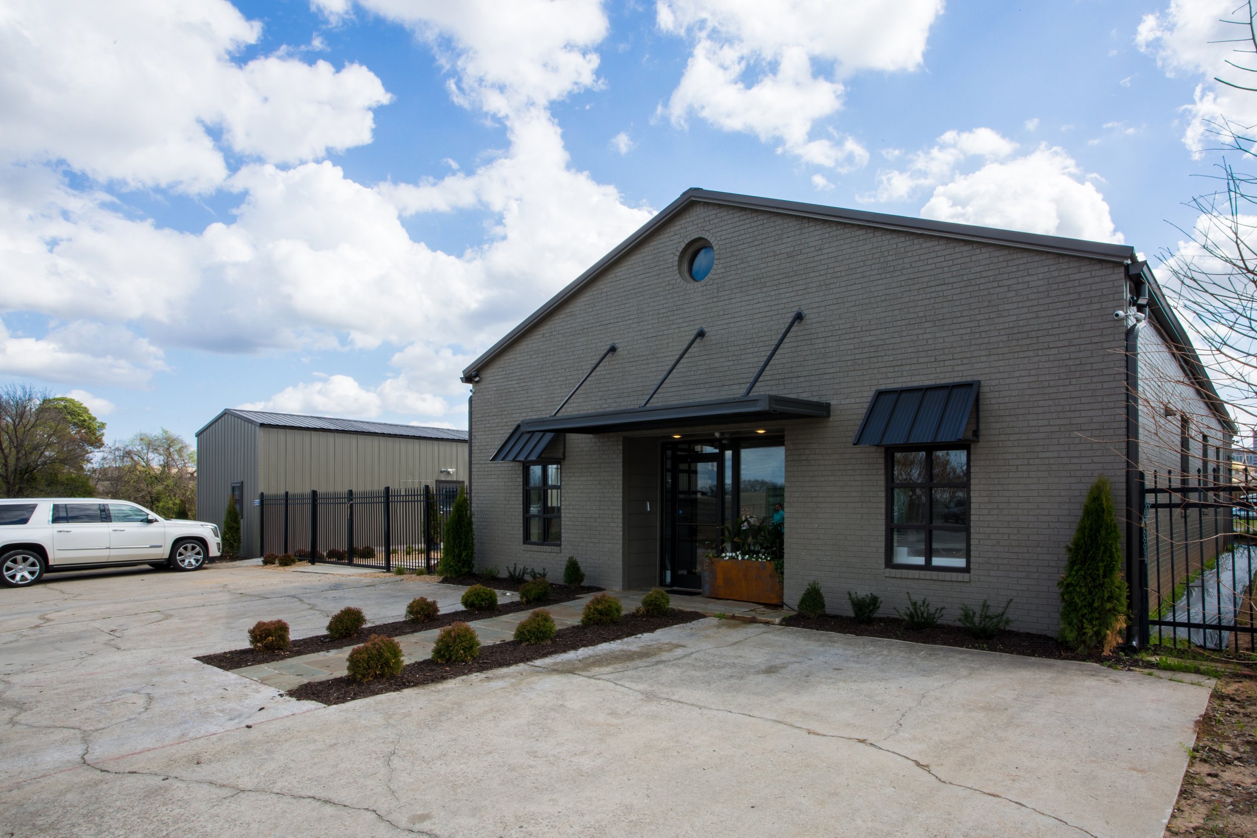 Modern gray brick building with black metal awnings, glass entrance, and round window in a residential or commercial area. Parked white SUV and small landscaped bushes and trees in the foreground under a partly cloudy sky.
