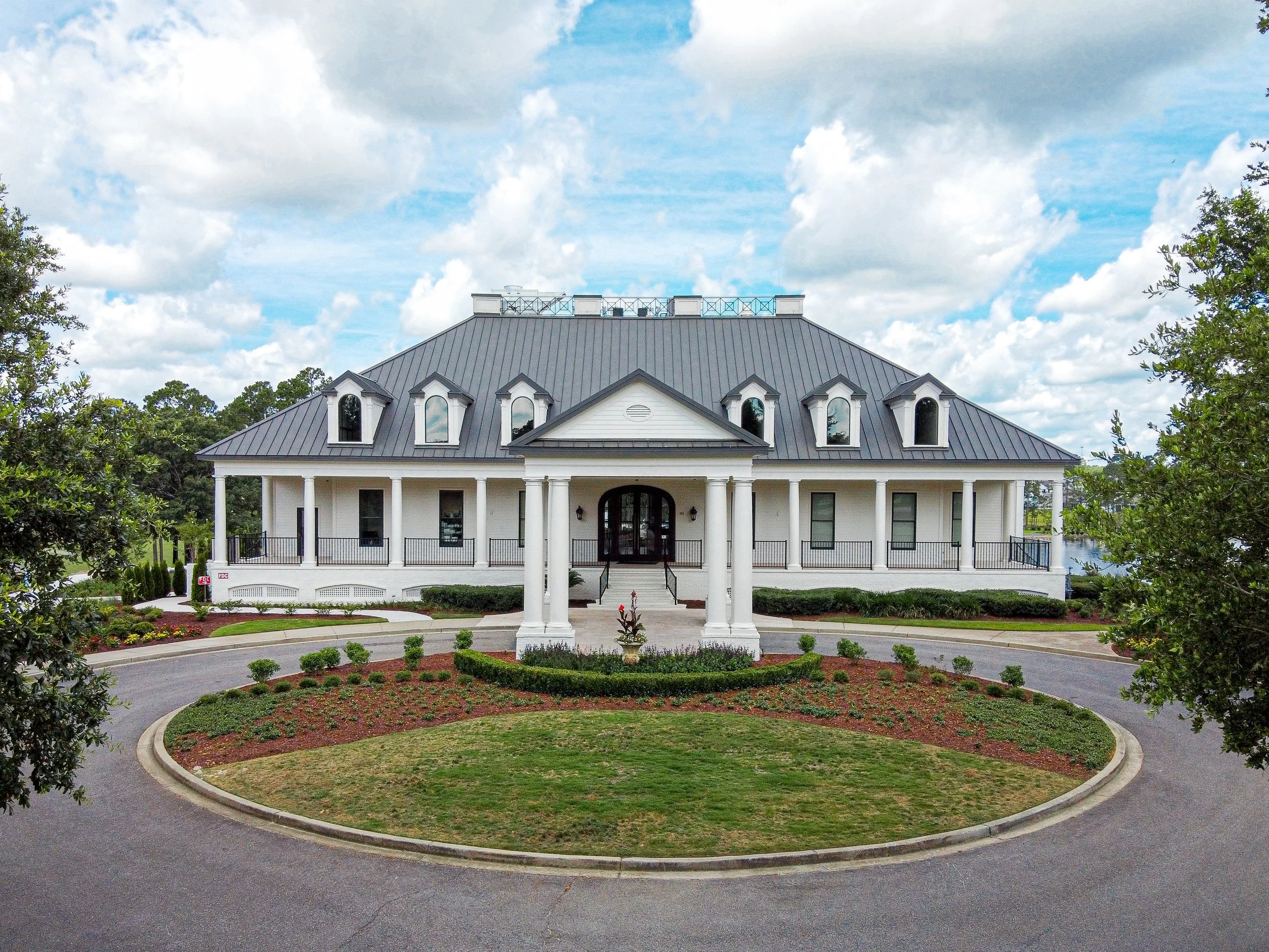 Large white house with a gray metal roof, multiple dormer windows, and a front porch with columns. The house is surrounded by a landscaped circular driveway with flower beds and greenery. The sky is partly cloudy.