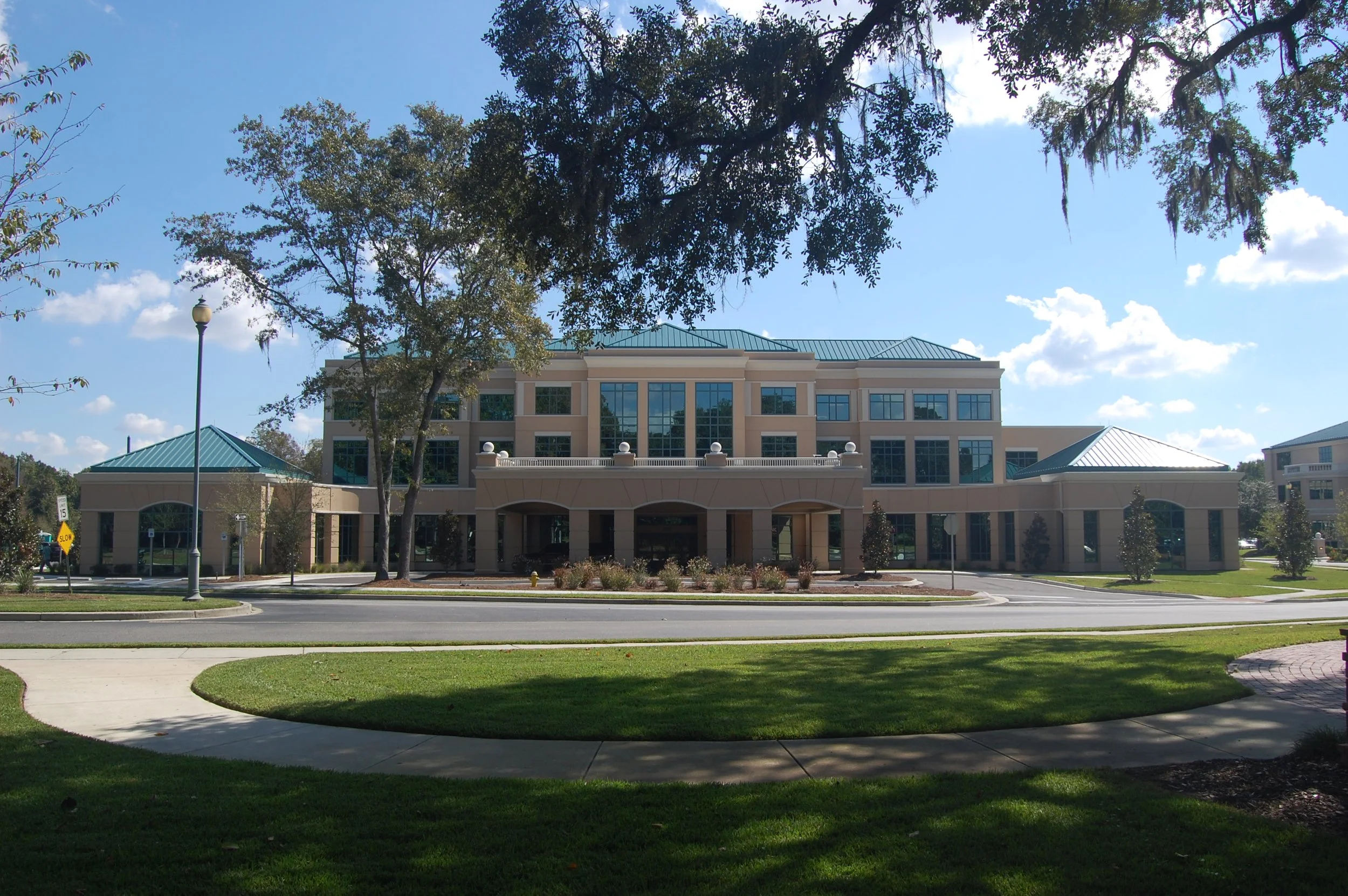 A modern, three-story building with large glass windows and a teal metal roof, surrounded by trees, a lawn, and a paved walkway, under a partly cloudy sky.
