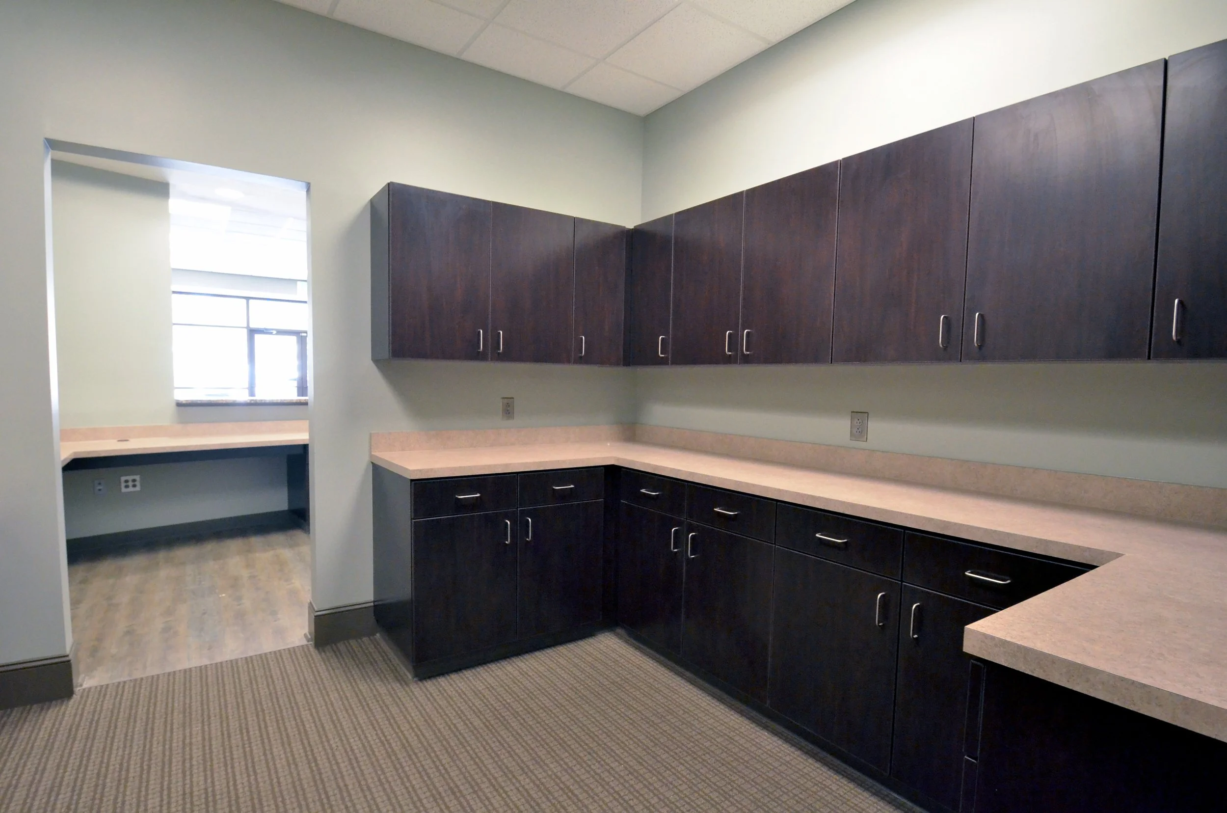 An empty office kitchenette with dark wood cabinets, light beige countertops, and a window with blinds in the background.