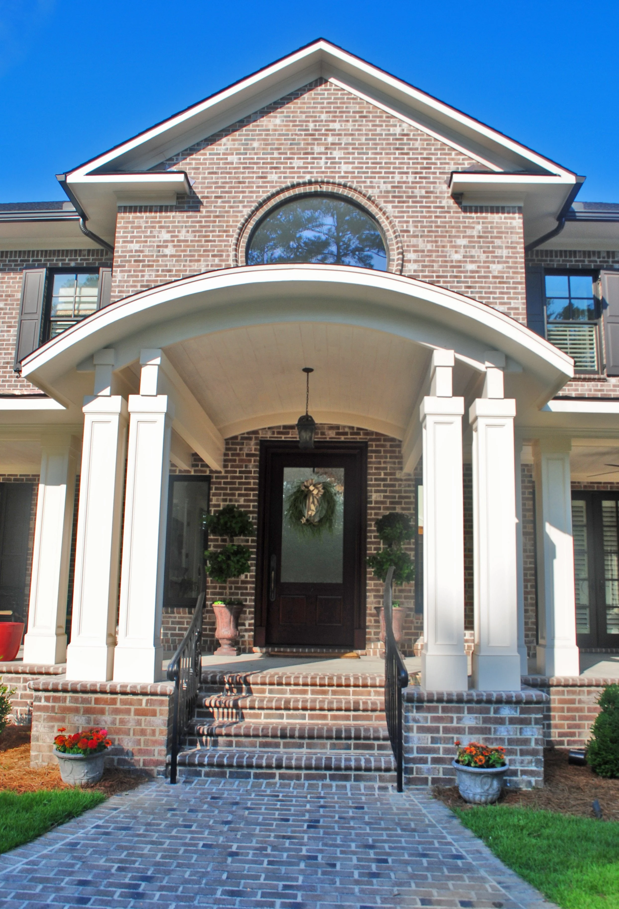 Front view of a brick house with a large arched window, white columns supporting the porch roof, and brick stairs with black railings leading to the black front door decorated with a wreath. Flower pots with red and orange flowers are on each side of