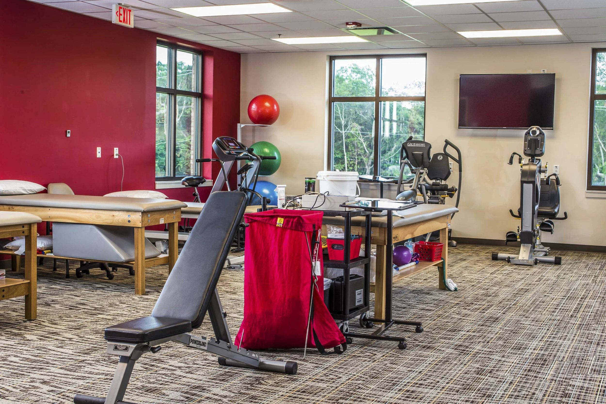 A therapy or rehabilitation room with various exercise equipment, including a treadmill, stationary bike, and elliptical machine, along with therapy tables, exercise balls, and a large wall-mounted TV, windows showing green trees outside.