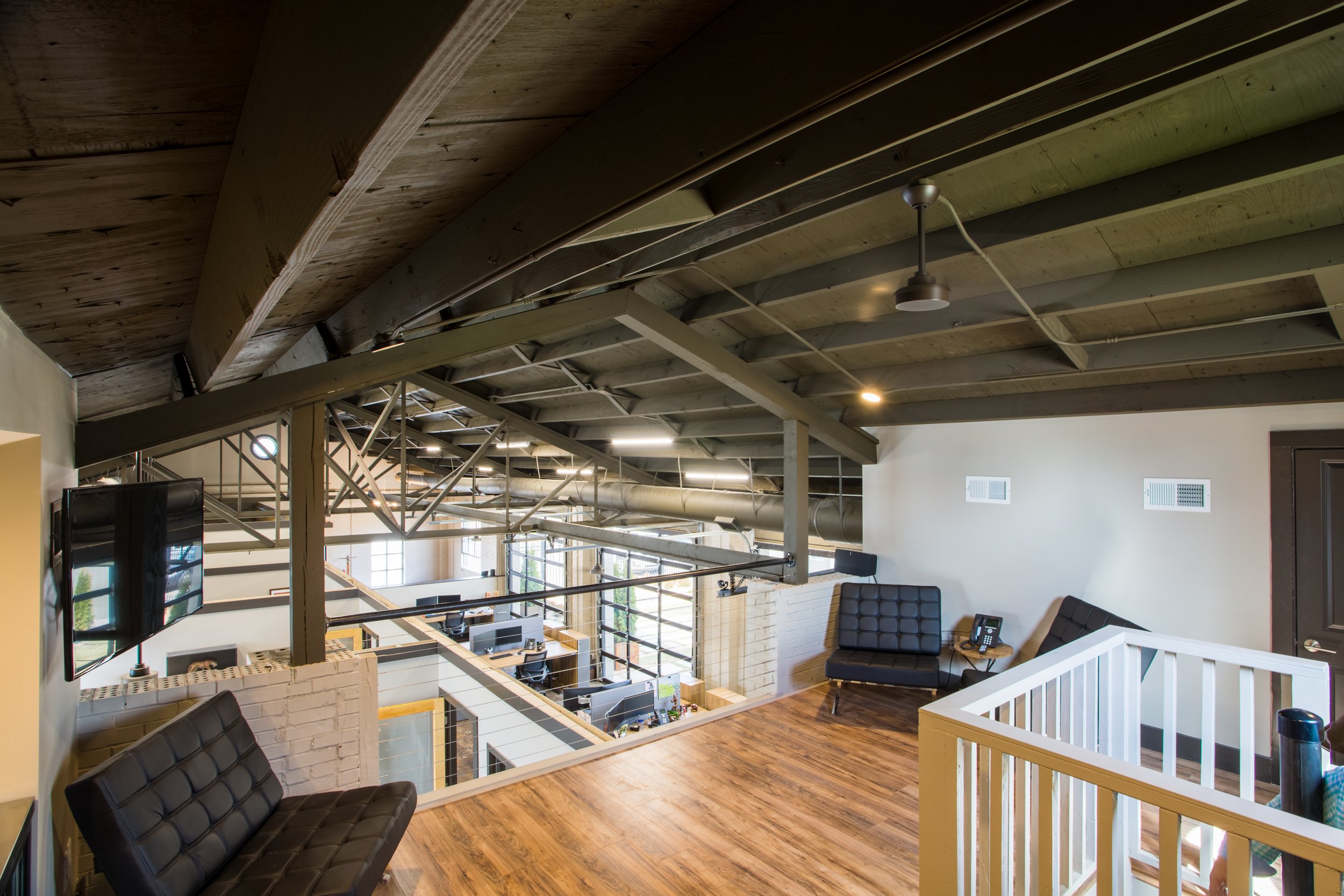 Interior view of an office space with exposed wooden ceiling beams, a TV on the wall, black leather seating, and large windows allowing natural light.