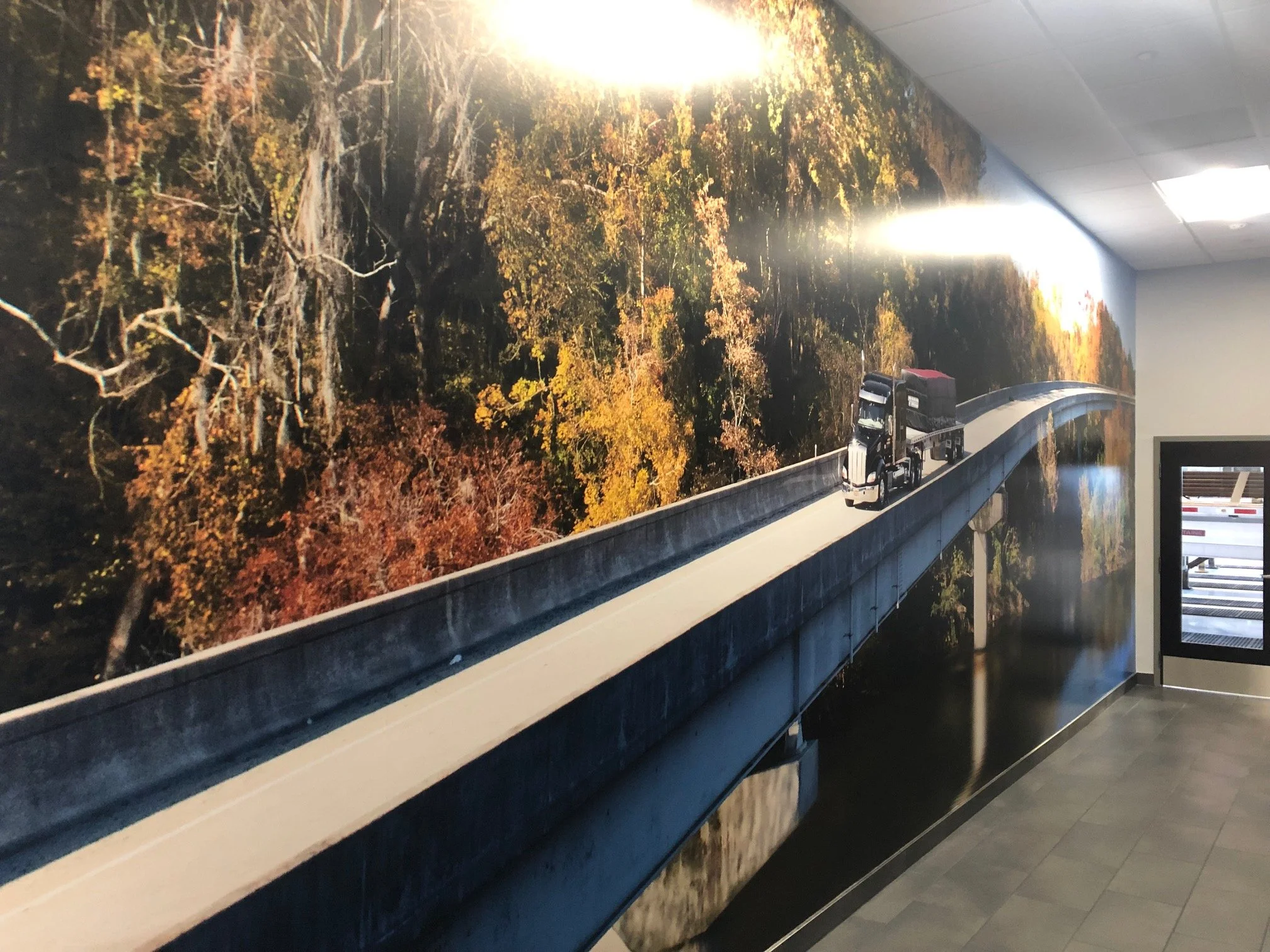 A wall mural of an autumn forest with trees displaying fall foliage in shades of yellow, orange, and brown. The mural features a highway with a semi-truck traveling on it, painted to blend into the natural scene.