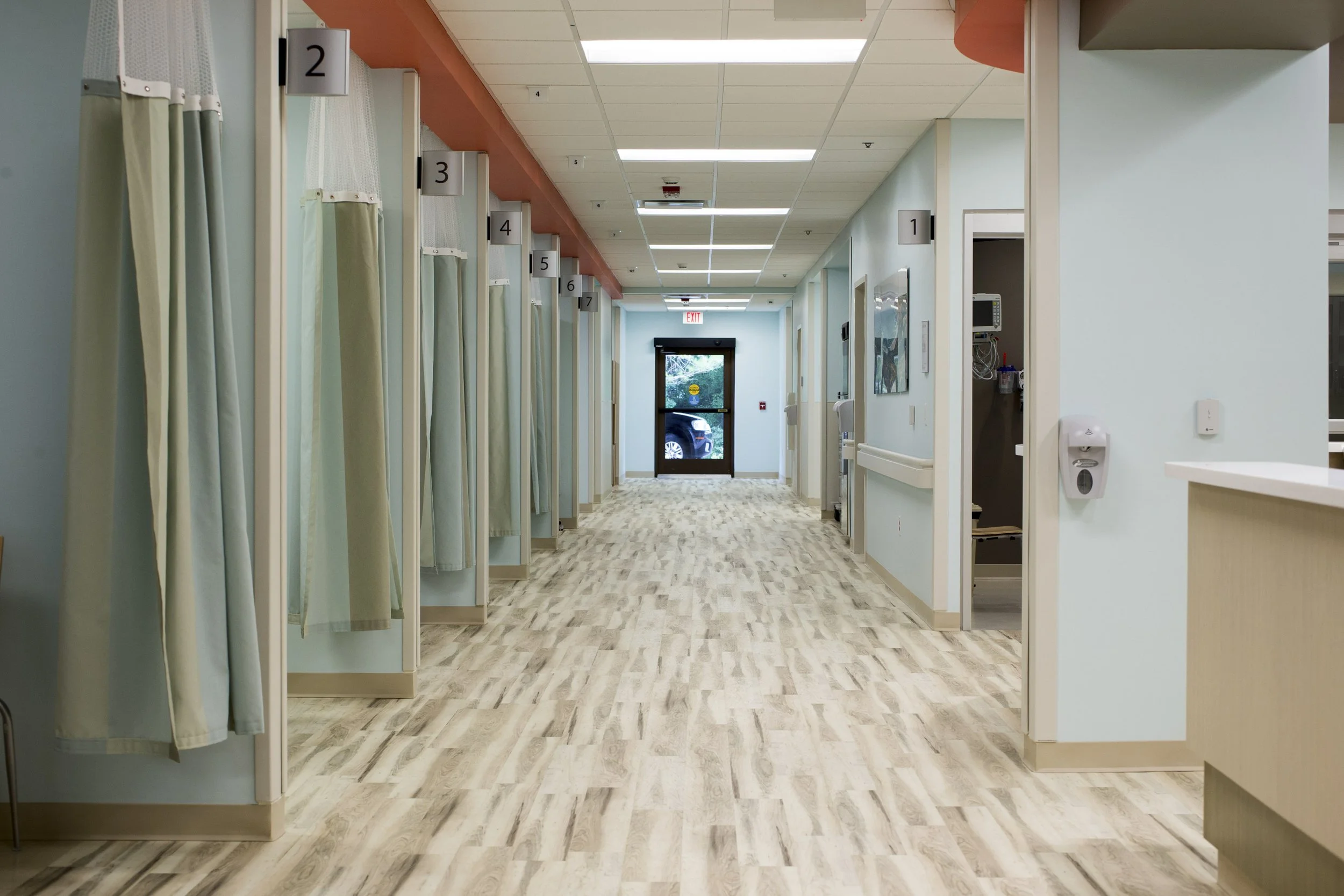 A hospital corridor with six patient rooms on the left, each with curtains and numbered signs above. The corridor has a light-colored wood floor, pale green walls, and a drop ceiling with fluorescent lighting. At the end of the hall, there is a glass