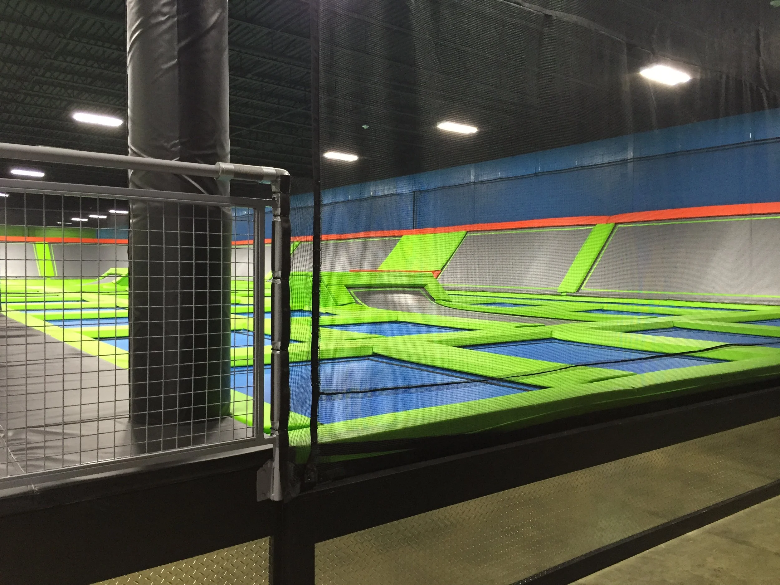 An indoor trampoline park with a large trampoline area enclosed by netting, and padded walls with vibrant green, blue, gray, and orange colors.