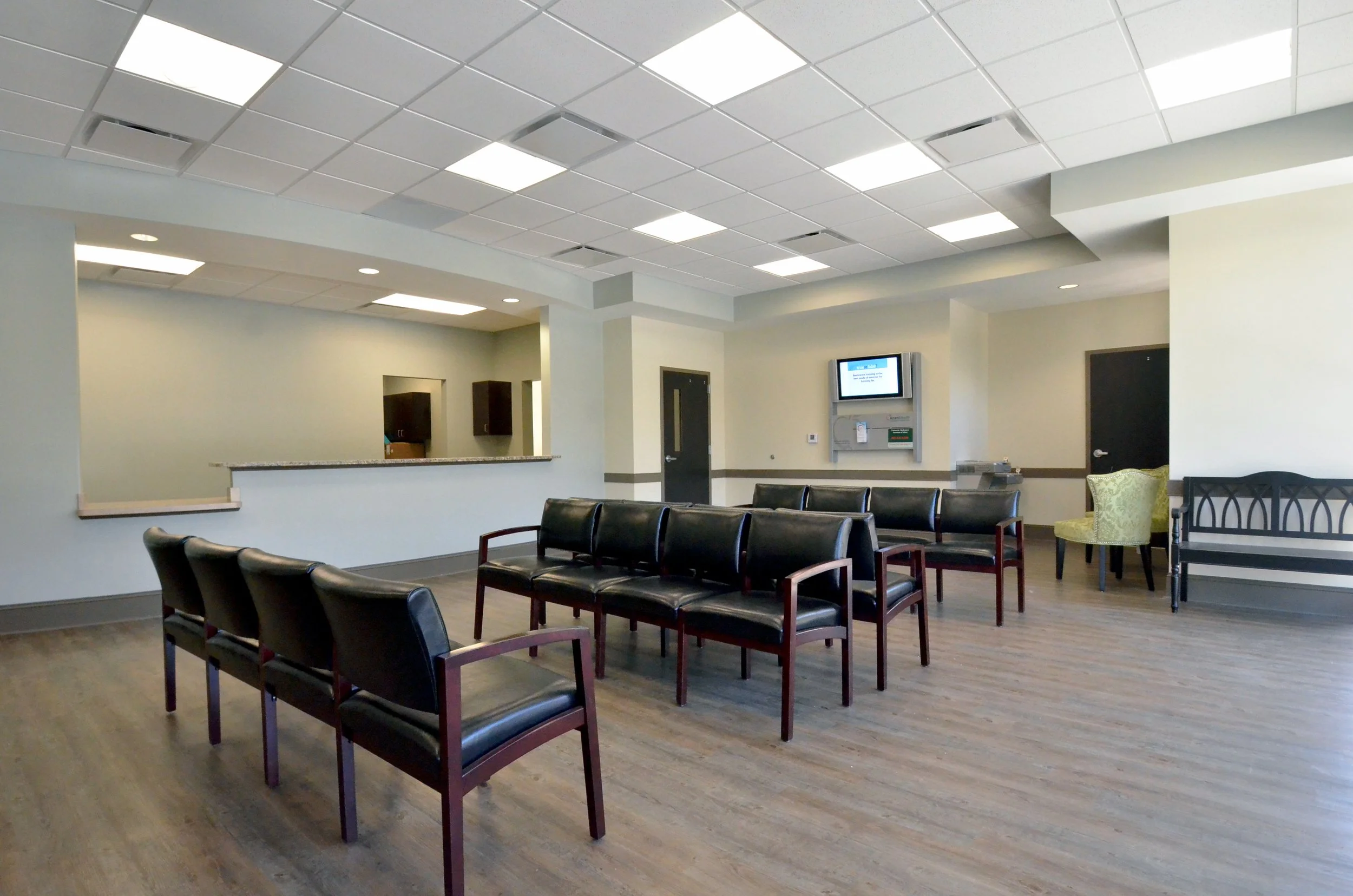 Waiting room with black chairs arranged in rows, a white wall with a television screen, and a kitchenette area in the background, in a medical office building.