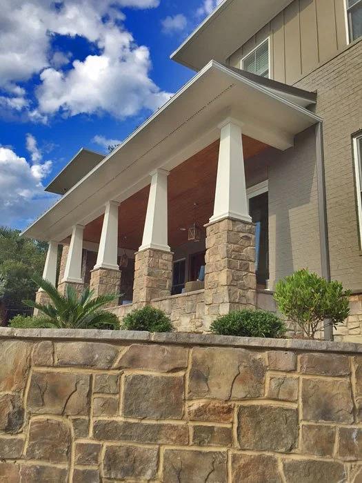 Front view of a house with a stone foundation, white columns supporting a covered porch, and a beige exterior with a second story. The porch has a wooden ceiling and the house is set against a partly cloudy blue sky.