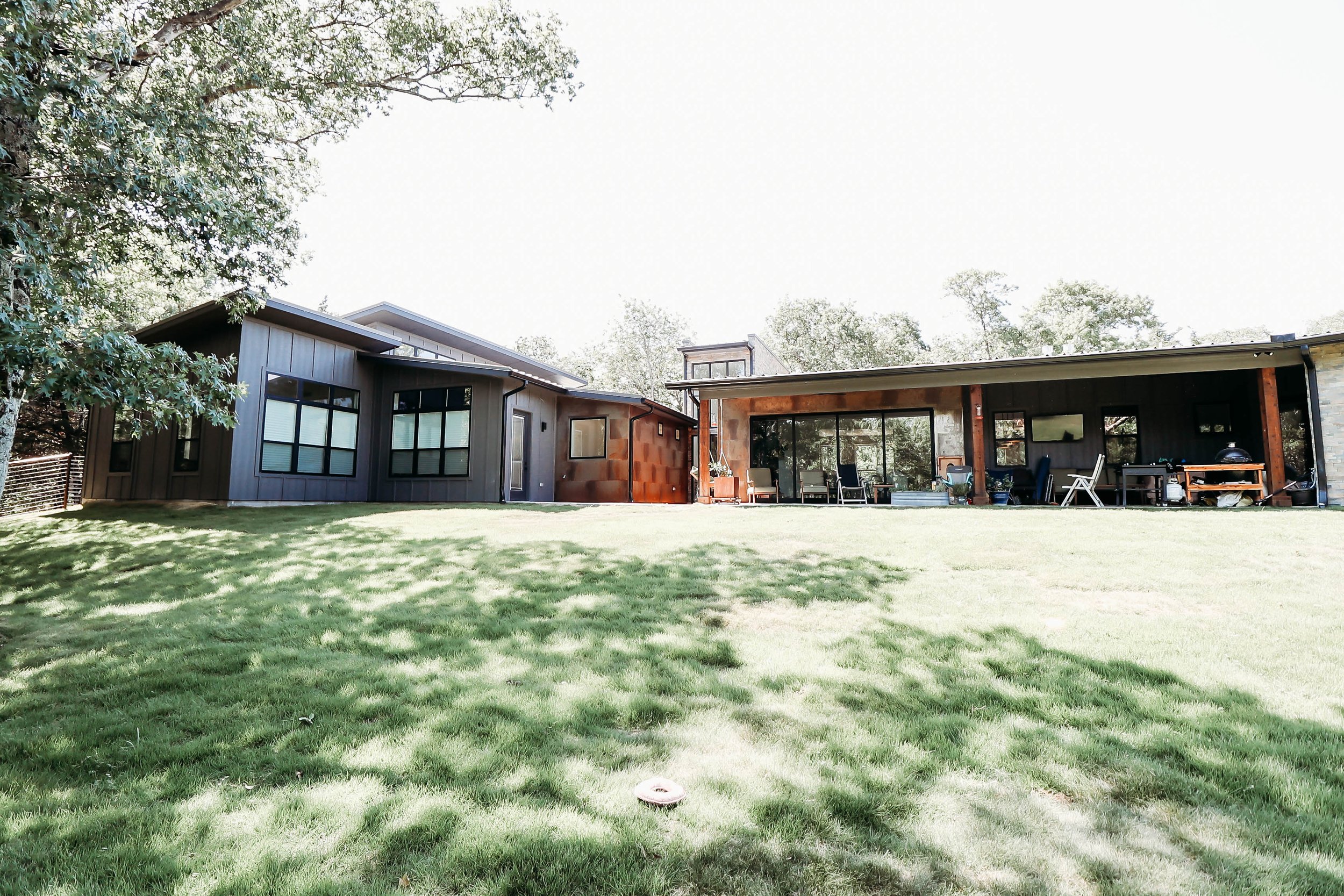 A backyard view with a well-manicured lawn, trees, and a modern house with large windows and an outdoor patio area with chairs and a grill.