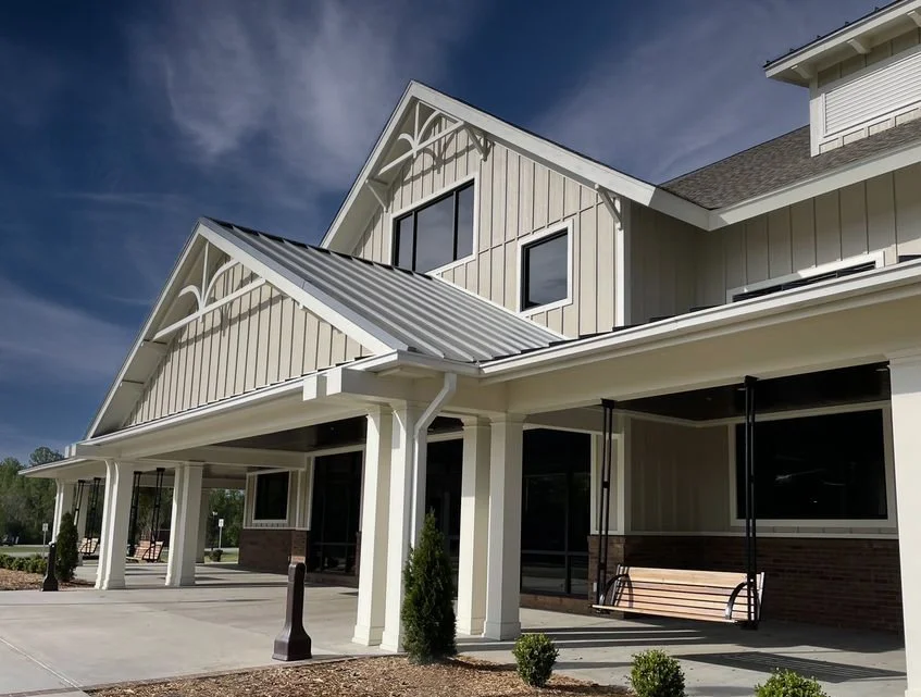 Exterior view of a modern multi-story building with white siding, large windows, and a metal roof, featuring a covered porch with a swing and small landscaping along the sidewalk.