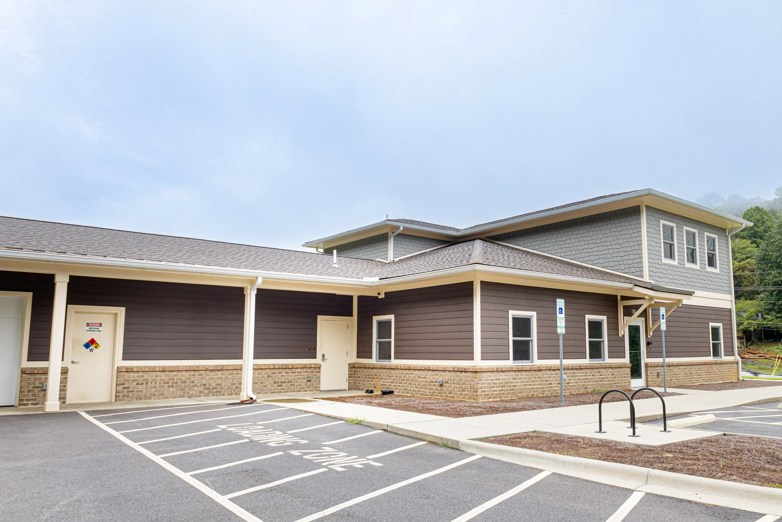 Exterior of a modern multi-story building with brown and gray siding, parking spaces, and handicap parking signs. Overcast sky in the background.