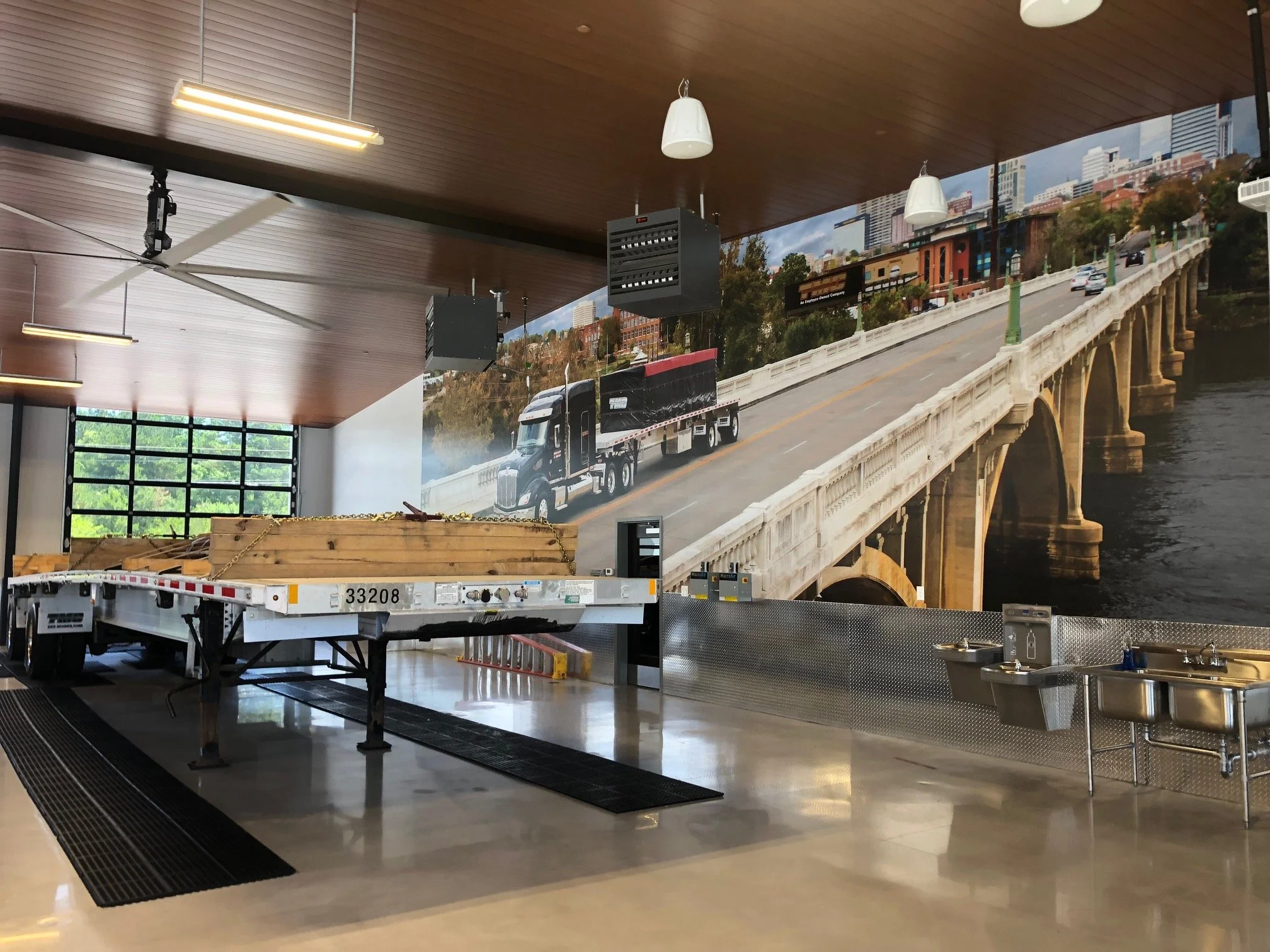 Indoor space with a wooden ceiling, ceiling fans, and lighting. There is a large mural or poster on the wall depicting a bridge over a river with cars, buildings, and trees in an urban setting. In the foreground, a flatbed truck with wood and chains is parked on a polished floor near a black mat.